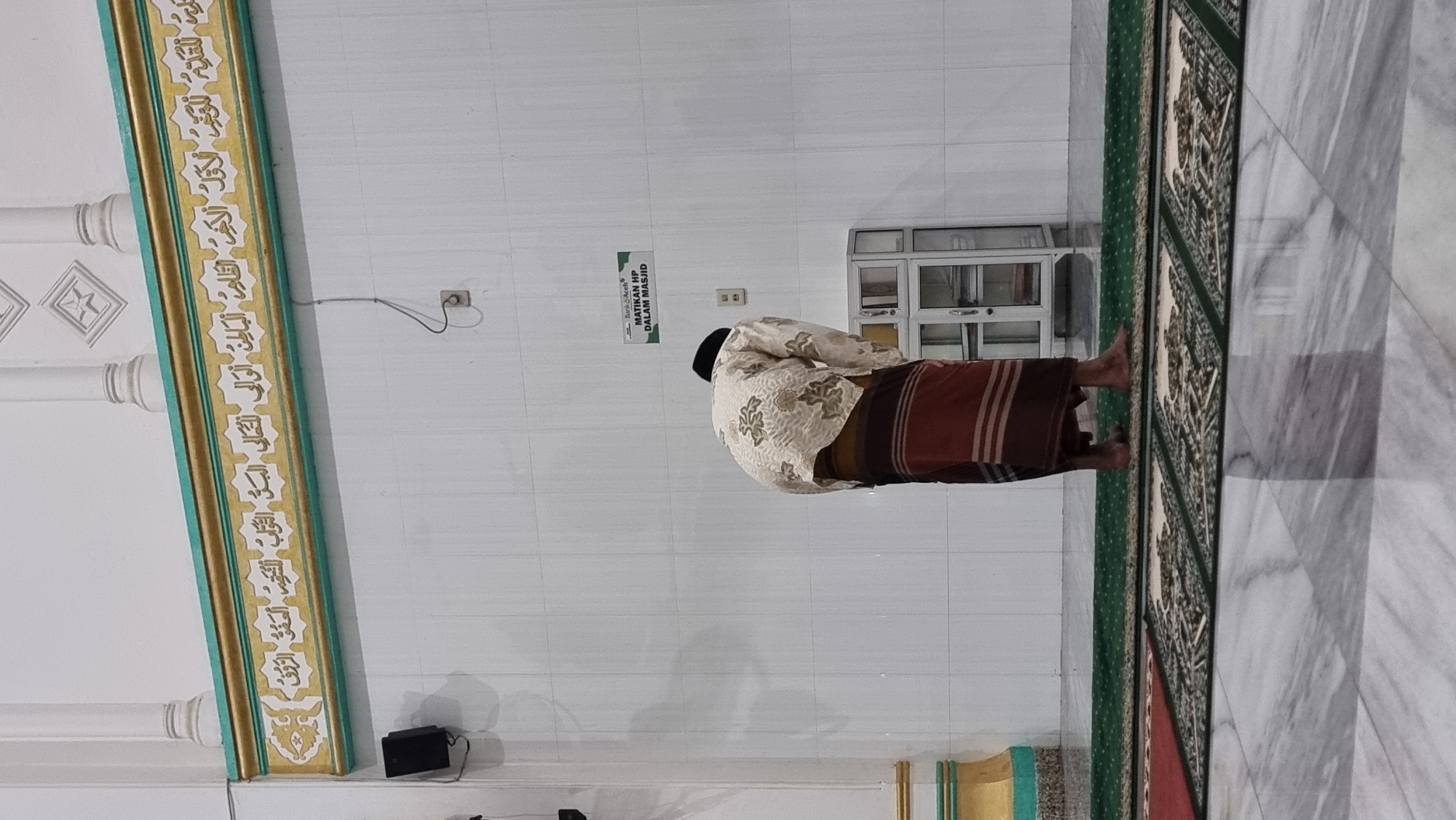 A man wearing a black peci, a patterned white shirt, and a brown sarong is bowing in prayer on a green prayer rug in a mosque. The mosque features white tiled walls, a green and gold decorative border with Arabic calligraphy, and white pillars. A sign on the wall reads "MATIKAN HP DALAM MASJID" (Turn off your phone in the mosque). A small white cabinet with glass doors is visible to the right of the man. The floor is made of polished marble. The image appears to be taken in Lapang Timu, Indonesia.