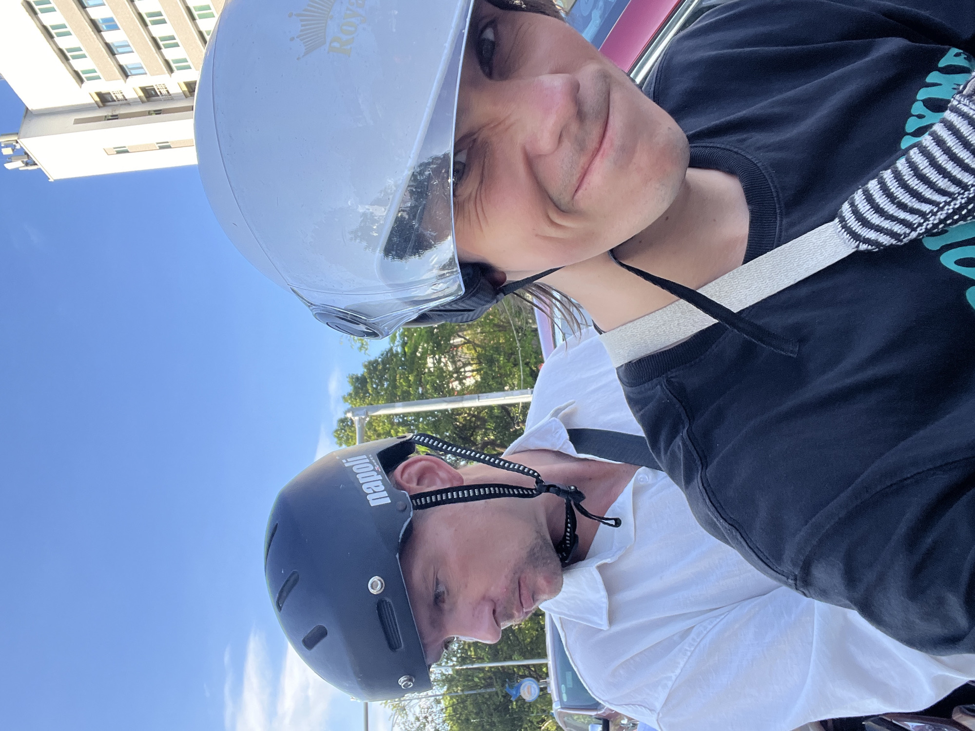 Two individuals are on a motorcycle in Đà Nẵng, Vietnam, with a white building and blue sky in the background. The person in the foreground, wearing a black t-shirt and a reflective silver helmet, is looking towards the camera with a playful expression, winking. The person behind them, wearing a white collared shirt and a black helmet, is looking to the left with a neutral expression. The image appears to be a selfie taken by the person in the front.
