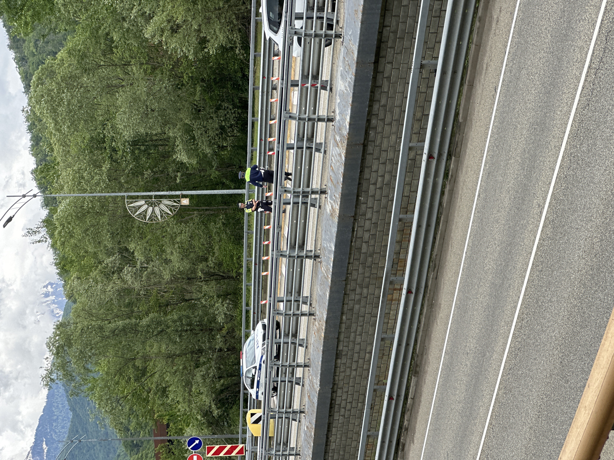 The image shows a bridge in Krasnaya Polyana, Russia. Two police officers stand on the bridge's pedestrian walkway, next to an unidentifiable white vehicle. One officer is wearing a dark blue jacket and the other a yellow reflective vest. Safety cones line the inside of the railing. Traffic signs are visible on the left. On the left side of the bridge is a tree-covered hillside. Mountains are visible in the background. The bridge spans a road with white lane markings. A decorative lamppost is seen on the side of the road.