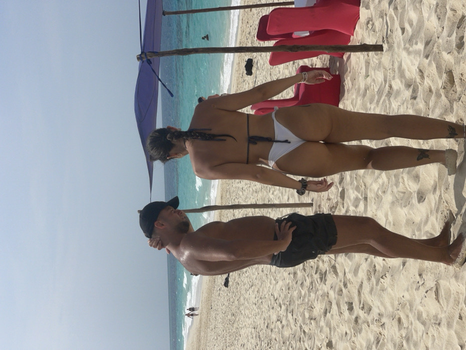 The image captures a sunny beach scene in Zanzibar, Tanzania. In the foreground, on light-colored sand, an adult male stands shirtless, wearing black swim shorts and a black baseball cap. He is positioned facing slightly left, with his head down and right hand on the back of his neck, his left hand on his hip. To his right, an adult female is seen from behind, wearing a white thong bikini bottom and a dark bikini top. Her dark hair is styled in a long braid, and she has butterfly tattoos on the back of each calf. Both individuals are barefoot, with sand on their feet. In the mid-ground to the right, a makeshift shade structure featuring a purple fabric canopy supported by wooden poles stands near the water's edge. Two red fabric-covered chairs are visible beneath the canopy. The turquoise ocean with small waves extends to the horizon under a clear, light blue sky. Distant figures include two individuals walking on the beach to the far left and at least two people swimming in the ocean to the mid-right. A dark object rests on the sand to the right of the walking figures.