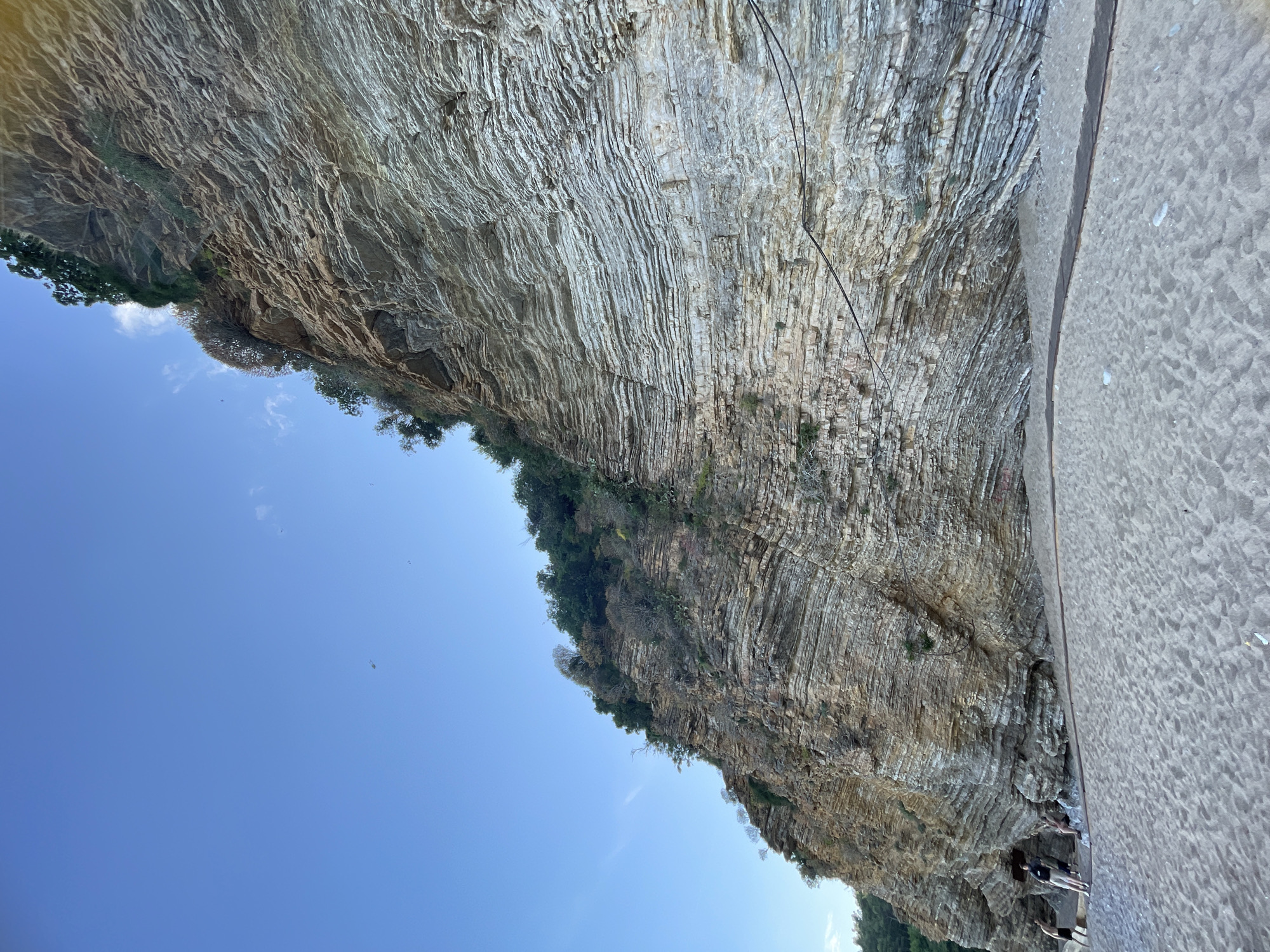 The image shows a large rock formation, possibly a cliff or mountain side, in an outdoor setting. The rock is layered with distinct striations, suggesting sedimentary rock or geological formations. Patches of vegetation, including trees and shrubs, grow on the rock surface.
In the lower right corner, there is a sandy area, possibly a beach or shoreline. A few people are visible in this area, and they appear to be standing or walking.
The sky is clear blue and the image is taken during daytime with good visibility.
The location is Budva, Montenegro.