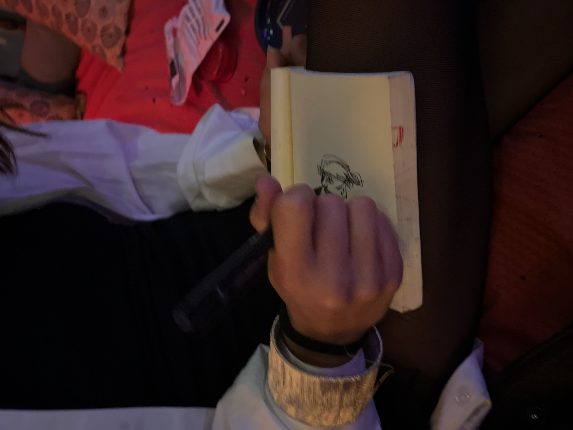The image captures an indoor scene, likely in the evening or at night, characterized by dim, warm, reddish-orange ambient lighting. The central focus is on a person's right hand, which holds a black pen or marker poised over a small, open, cream-paged sketchbook. On the left page of the sketchbook, there is a distinct, simple line drawing of a stylized head or face. Faint red markings are visible along the edge of the right page.
The person wears a large, metallic, textured cuff or bracelet on their wrist, positioned over the sleeve of a white shirt, which is visible beneath a darker garment, possibly a jacket or top. In the blurred background to the left, there are patterned cushions and reddish fabric, suggesting a bed or sofa. Further back, a crumpled white object with some indistinct black text and a red cup or container are partially visible. The scene depicts a quiet moment of creative activity, with the individual focused on sketching. There are no explicit visual cues within the image to indicate the provided location context of Barcelona, Spain.