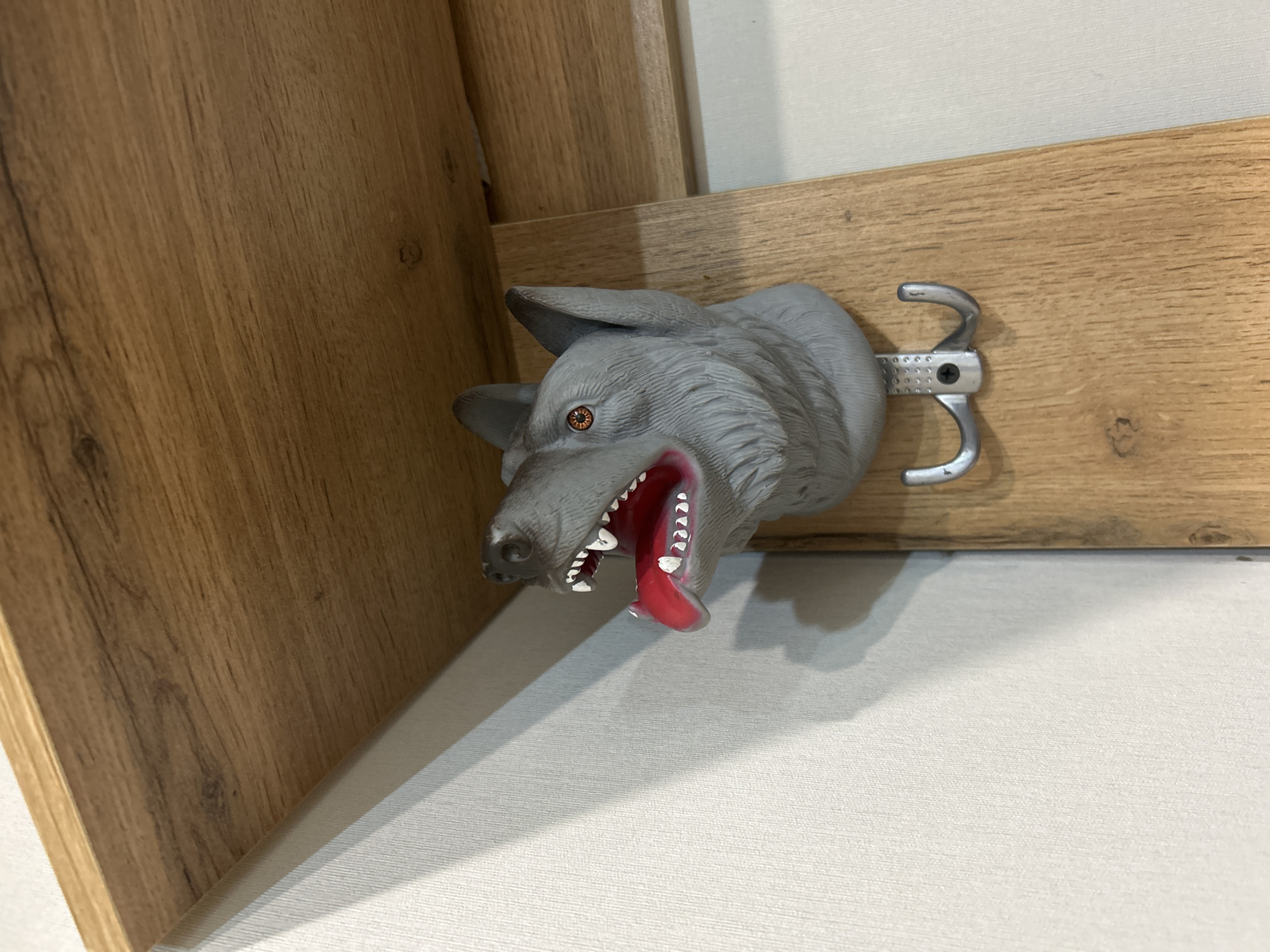 A gray wolf head sculpture with an open mouth and protruding red tongue is mounted as a decorative hook on a light-colored wooden board attached to a wall. The hook is made of silver metal. The context suggests an interior space, with wooden paneling on the left and a white wall behind. There is no evidence to place this image in the city of Уральское, Russia, or to provide any specific location details. There are no people visible in the frame.