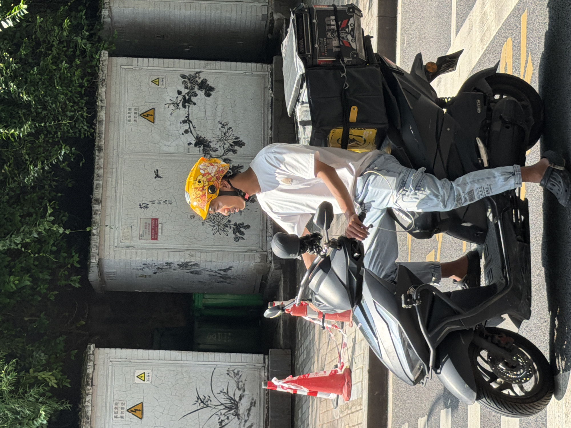 In Yuzhong District, China, a young man wearing a yellow helmet decorated with cartoon characters rides a black scooter. He is dressed in a white t-shirt and light blue cargo pants. The scooter has a large black insulated box attached to the rear, likely for food delivery, with yellow accents and various stickers and labels. The man is looking to his right, and his left hand is on the scooter's handlebar. The scooter is stopped at a crosswalk, indicated by yellow lines on the asphalt. In the background, there are two utility boxes with Chinese writing and warning signs, one featuring a yellow triangle with a lightning bolt symbol. Green trash bins are visible between the utility boxes. Orange traffic cones, one partially wrapped in red and white tape, are placed on the sidewalk to the left. Lush green foliage hangs over the utility boxes and the top of the frame. The scene appears to be a typical urban street environment on a bright, sunny day.