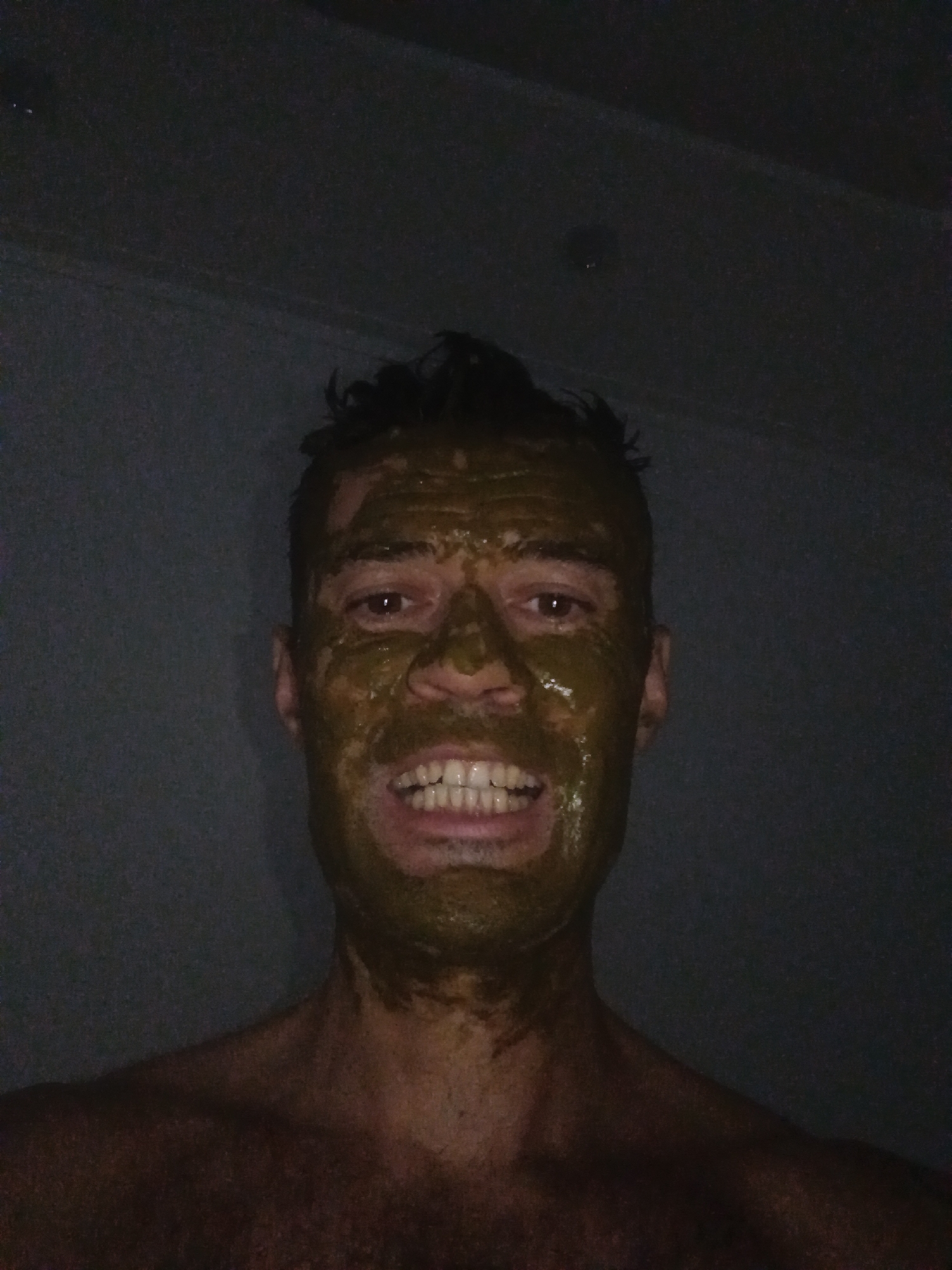 A medium shot depicts a man with dark hair and skin covered in a thick, dark green substance, likely mud or a cosmetic clay mask. He faces the camera with a wide, toothy grin. The background is a dark, mottled gray, possibly a wall. The lighting is dim, creating a shadowy effect. The man is shirtless. There's no specific evidence of the location being Mũi Né, Vietnam.
