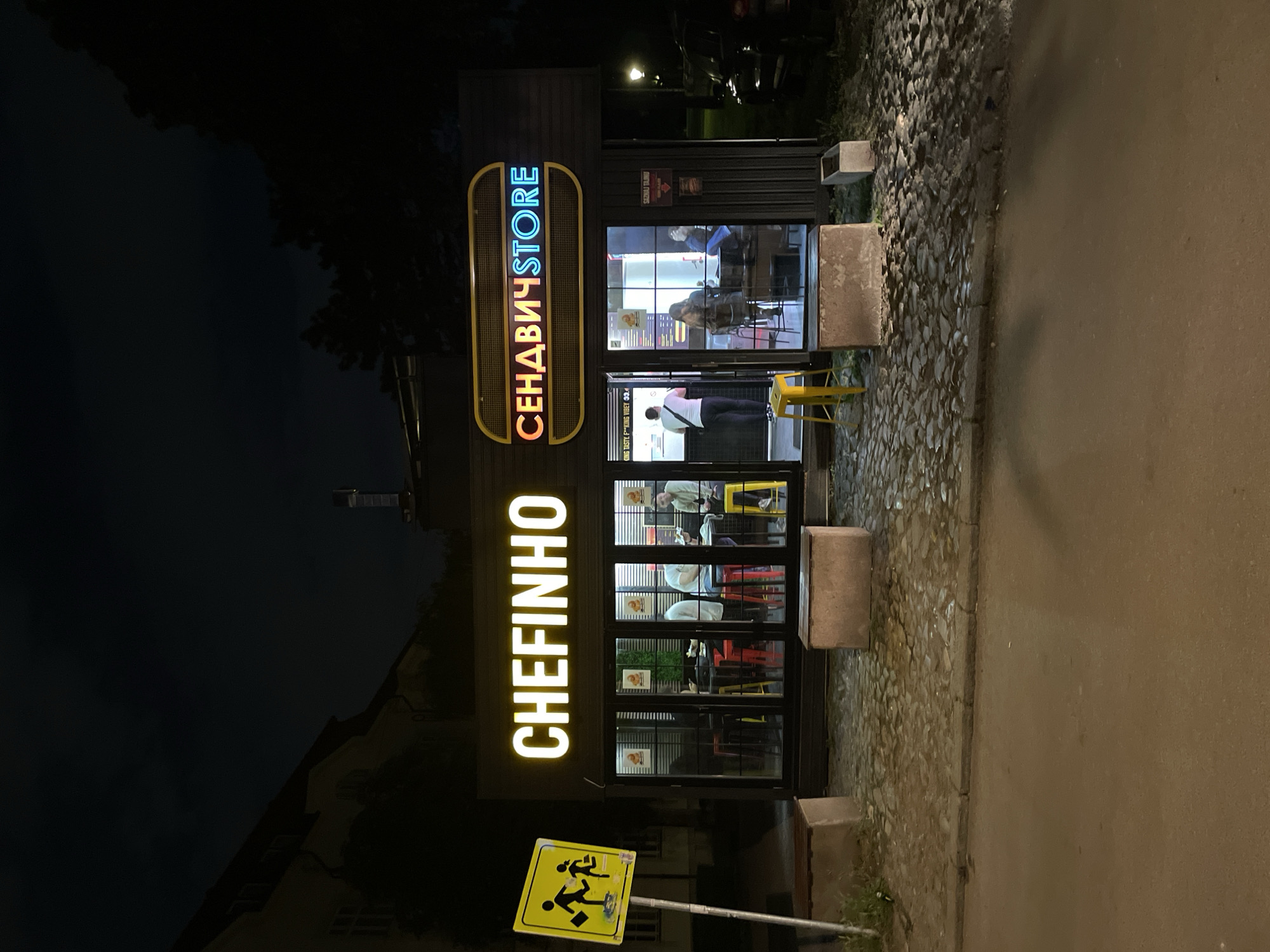 The image shows the facade of a building identified as "CHEFINHO CЕHДABИЧSTORE" located in Zemun Urban Municipality, Serbia. The name "CHEFINHO" is vertically aligned and lit with yellow neon. Above it, "CЕHДABИЧSTORE" is lit with multi-colored neon and horizontally aligned. The building has multiple windows, with some people seen inside. A yellow road sign is present. The building is beside a path.