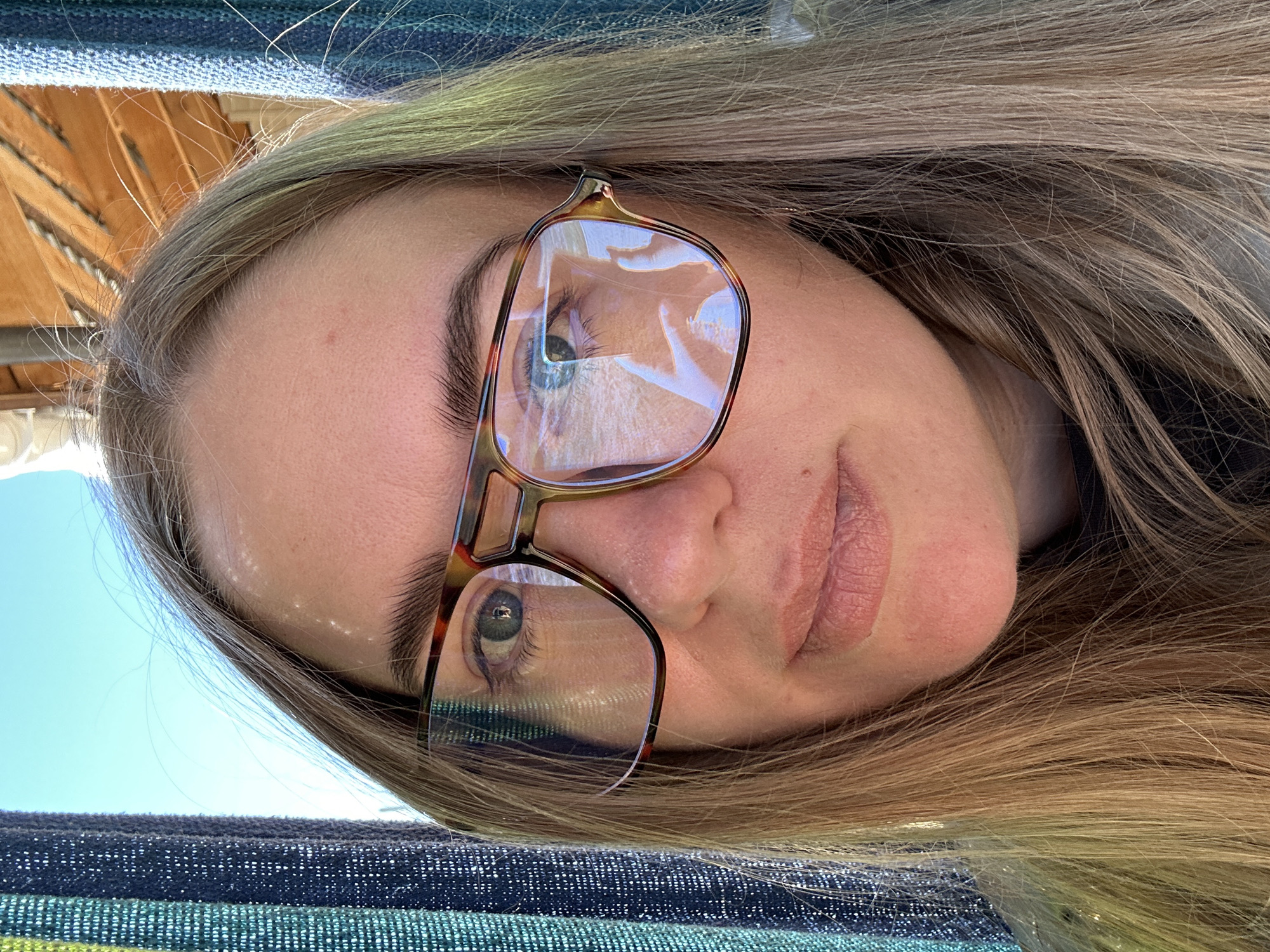 A close-up, upward-angled selfie shows a woman with long, light brown hair wearing tortoiseshell-patterned glasses. Her face is centered in the frame, with her nose and mouth clearly visible. Her right eye is visible through the glasses, with the lens reflecting the sky and possibly a structure. The left eye is partially obscured by the glasses. The background includes a striped fabric and hints of a wooden structure. The image does not contain sufficient information to determine the location of Уральское, Russia, or any actions, interactions, or context beyond what is visible in the close-up selfie.