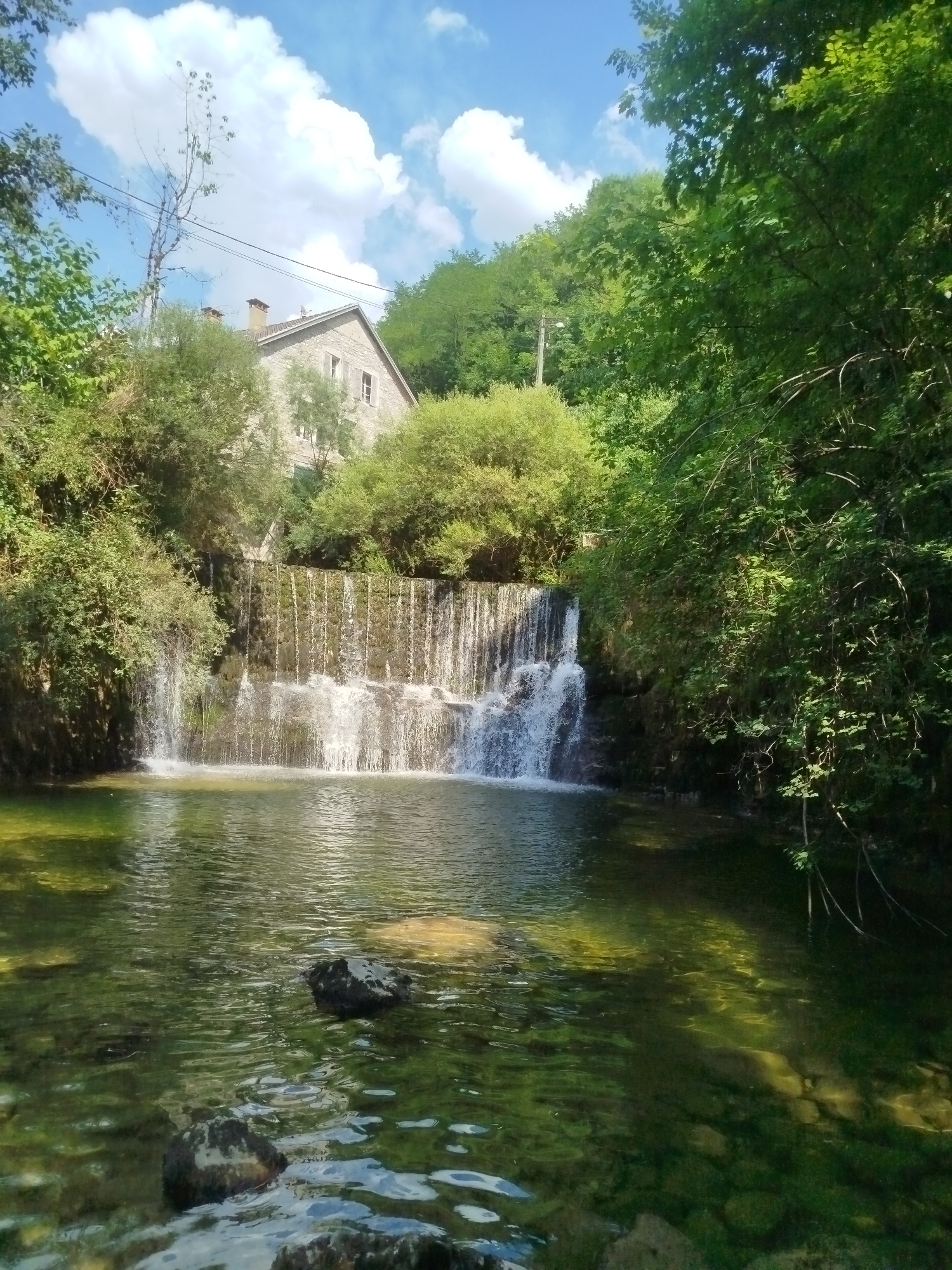 The image depicts a scenic view of a waterfall cascading into a shallow, clear body of water. The water is partially obscured by large, dark rocks protruding from its surface. Lush green trees and dense foliage frame the waterfall on both sides, with a stone building situated on a higher elevation behind the trees. The sky above is a vibrant blue, dotted with fluffy white clouds. The presence of overhead wires and a utility pole suggests that the location is within or near an inhabited area. While the precise location in Saint-Claude, France, is not explicitly identified, the scene suggests a natural setting within or on the outskirts of a settlement. There are no people or animals visible in the image.