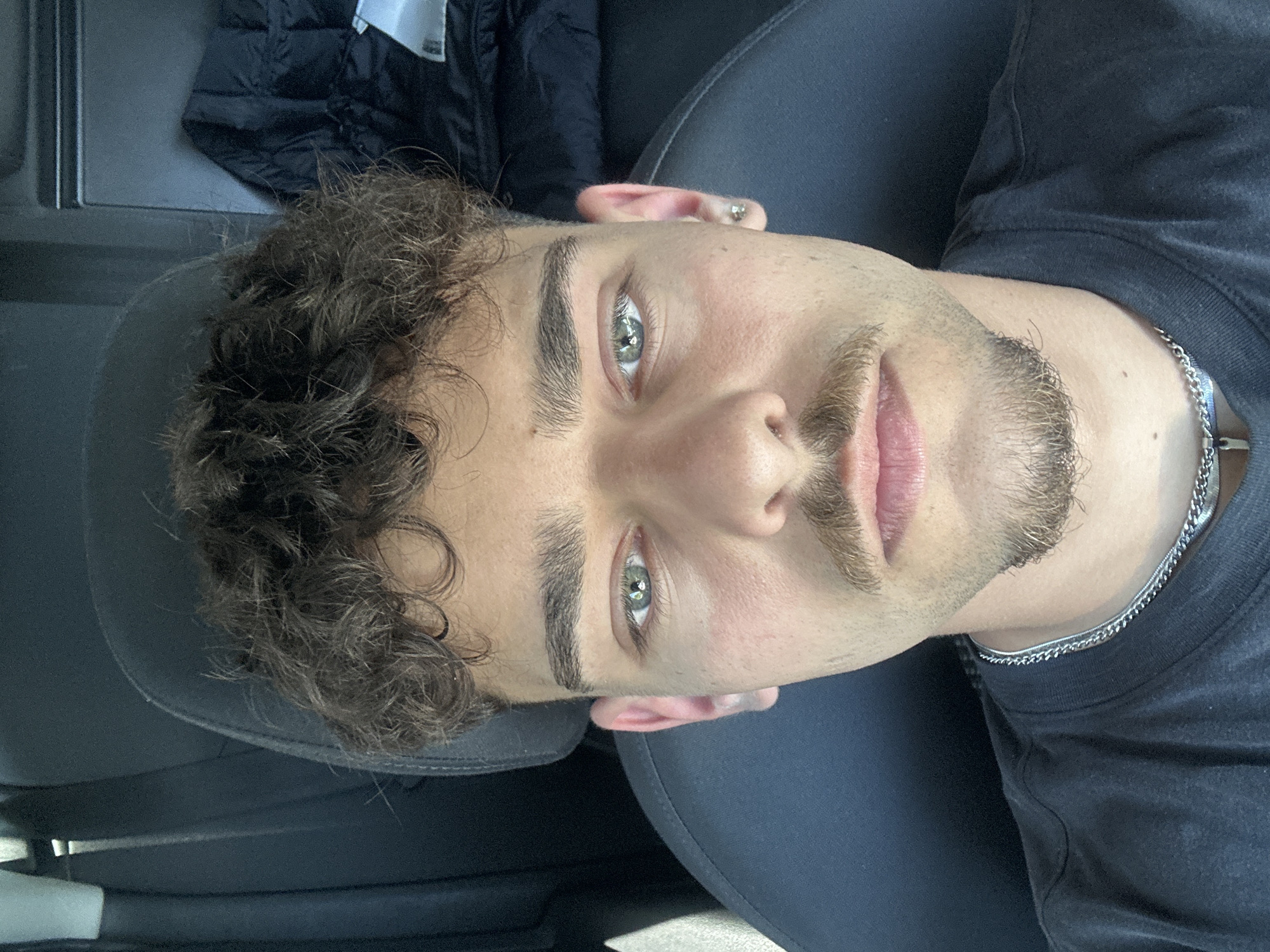 A selfie-style photograph depicts a young man with light skin, light brown curly hair, and stubble. He has light-colored eyes. He wears a dark t-shirt and a silver chain necklace. He appears to be seated in a car, as evidenced by the car seat and interior trim visible. A jacket is visible in the upper left. The photo is taken at an angle, likely from above. The location of the image, Ravnishte, Bulgaria, is only known because of the prompt instructions and is not visually supported by the image.