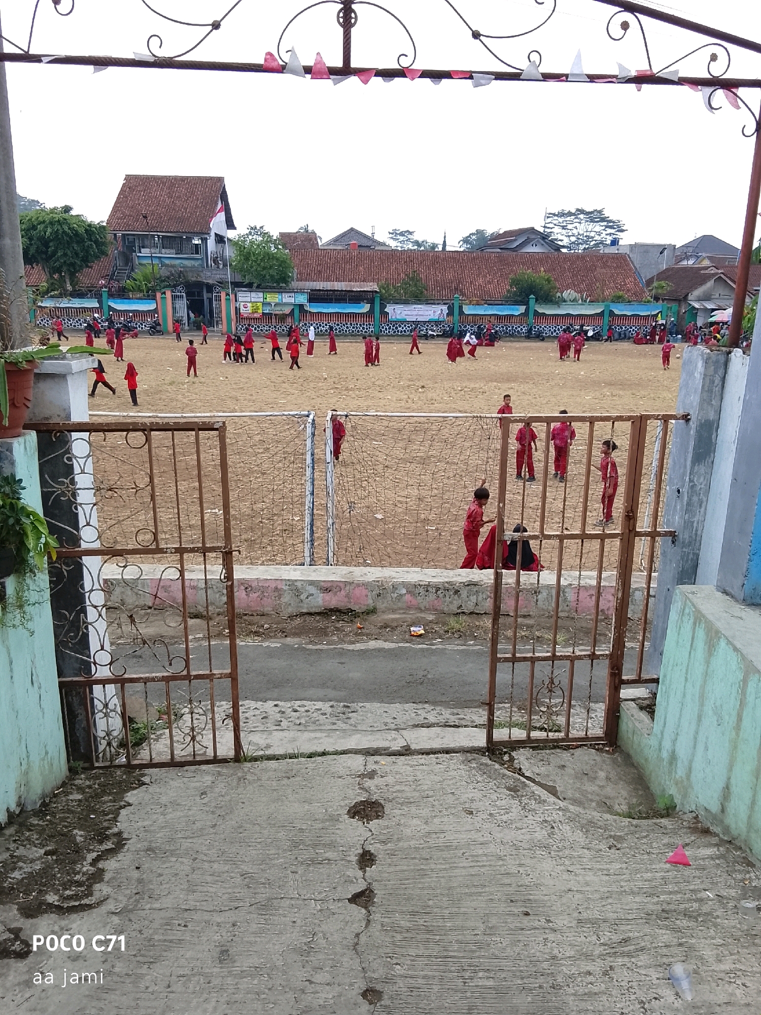 The image shows an outdoor scene with children playing on a dirt field. In the background, there are several buildings with red tile roofs, some appearing to be school buildings. A flagpole with an Indonesian flag is visible. The foreground features a rusty metal gate and a concrete pathway. The sky is overcast. Several children, most of whom are wearing red uniforms, are scattered across the field, engaged in various activities, possibly playing a game. The scene appears to be a schoolyard in the city of Tasikmalaya, Indonesia, given the presence of the Indonesian flag and the architectural style of the buildings, which are common in that region.