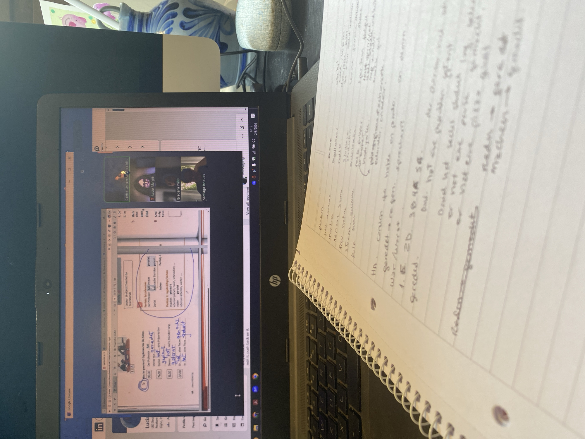 A person is participating in a virtual meeting on a laptop, possibly a class or seminar, based on the visible content on the screen. The meeting involves multiple participants. The laptop is placed on a desk with a notebook. The notebook contains handwritten notes in German. The laptop screen displays a PowerPoint presentation in German, which appears to be related to German grammar and vocabulary. The meeting is being conducted through an online platform. The name of the meeting host/institution is "Centro Universitario de Idi," and the date displayed on the taskbar is 2/5/2026. No specific information about the location in Villaviciosa, Spain, is provided.