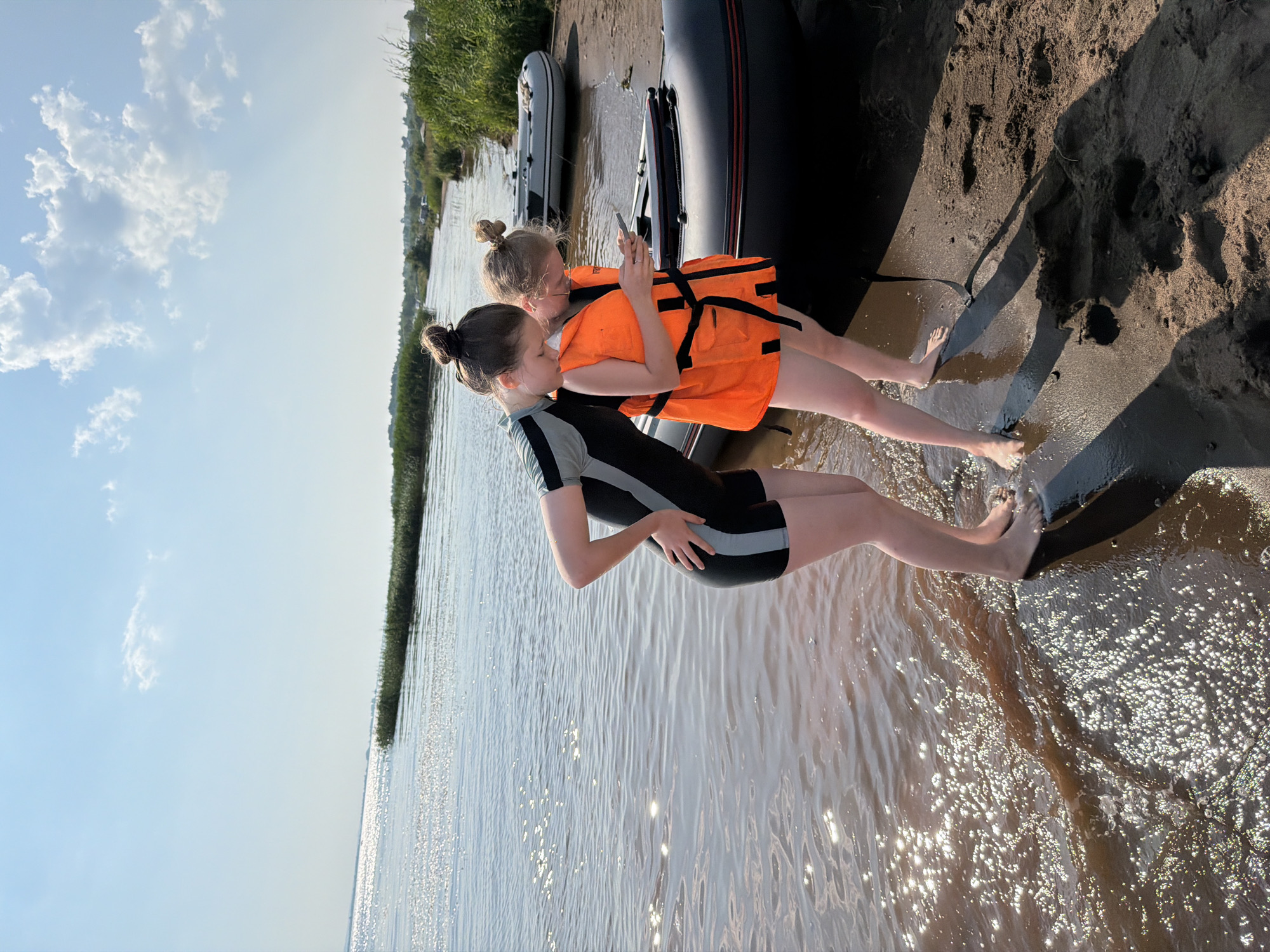 Two young females, one wearing a black and gray wetsuit and the other wearing an orange life vest over a bathing suit, stand on a sandy shore beside a dark-colored inflatable boat. The female in the wetsuit is leaning forward, looking down at the water. The female in the life vest is holding a smartphone and appears to be looking at it. Another lighter-colored inflatable boat is visible further up the shore, and the background shows a body of water, reeds, and trees under a bright, cloudy sky. The scene is consistent with a recreational area near the Neva River in the vicinity of Krasny Bor, Leningrad Oblast, Russia, a location known for its natural beauty and outdoor activities.