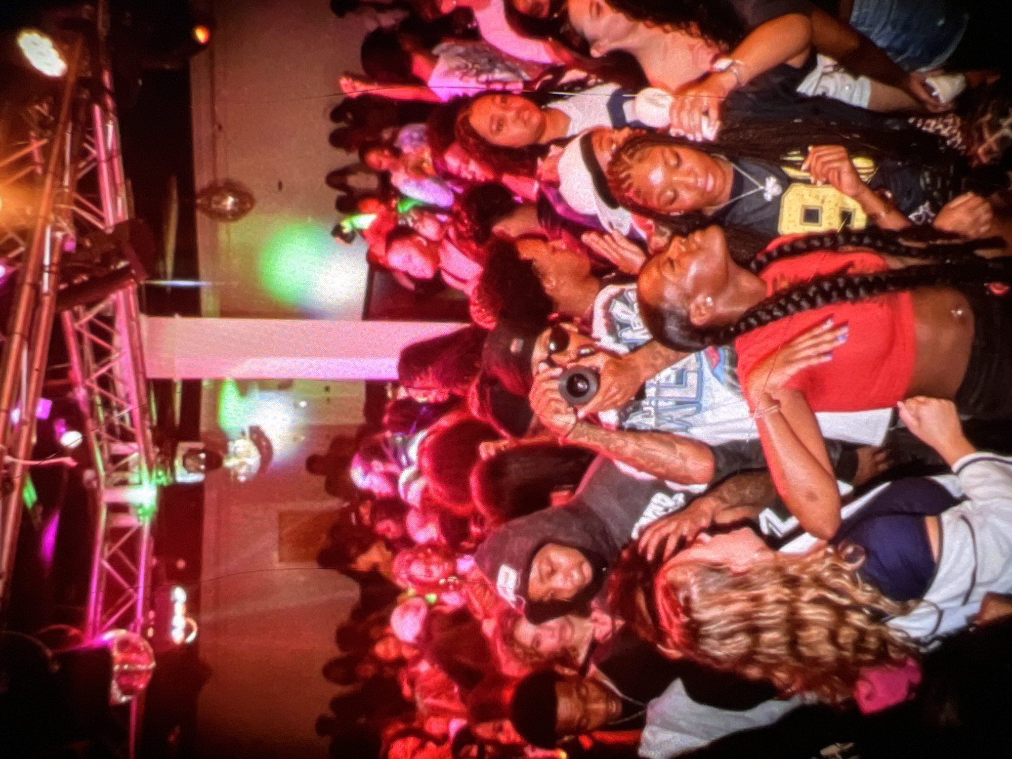 The image captures a lively indoor gathering, possibly a club or party, in Lagos, Nigeria. A crowd of young adults is visible, some engaged in dancing and socializing. The atmosphere is vibrant with colorful lighting, including pink and green hues, illuminating the space. Several individuals are holding drinks and phones, with one person in the foreground appearing to take a photograph with a camera. The scene is crowded, with people in close proximity, suggesting a dynamic social event. The background features disco balls and stage lighting, indicative of a venue designed for entertainment.