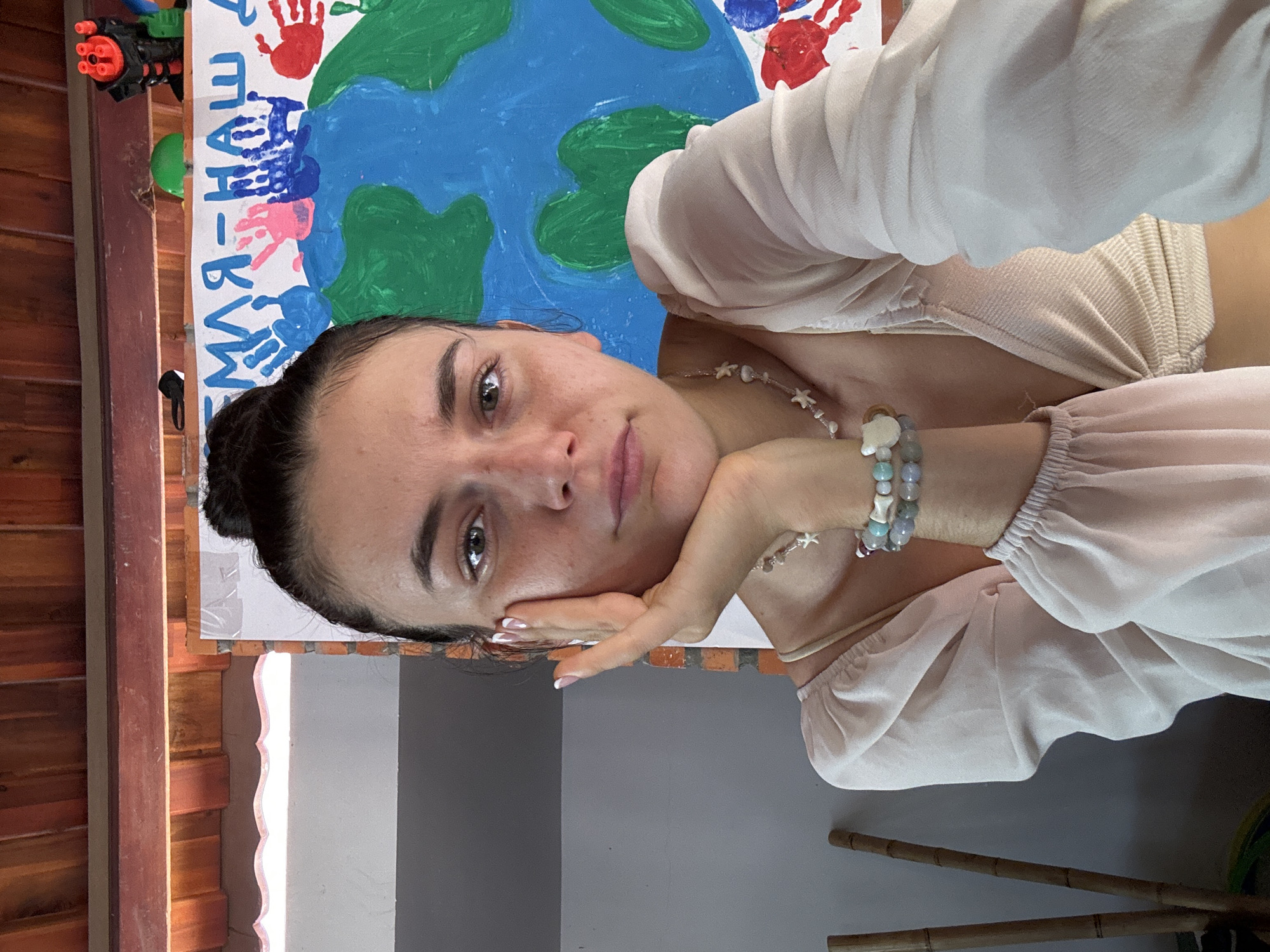 A selfie shows a woman with brown hair pulled back in a bun and light skin, resting her chin on her hand, wearing a light-colored blouse and beaded jewelry. Behind her is a child's artwork depicting the Earth with handprints in various colors and handwritten text in a language other than English. In the background, there's a wooden wall and a glimpse of a room interior with a gray wall and bamboo objects. It is impossible to determine the specific location in Mũi Né, Vietnam, from the given image.
