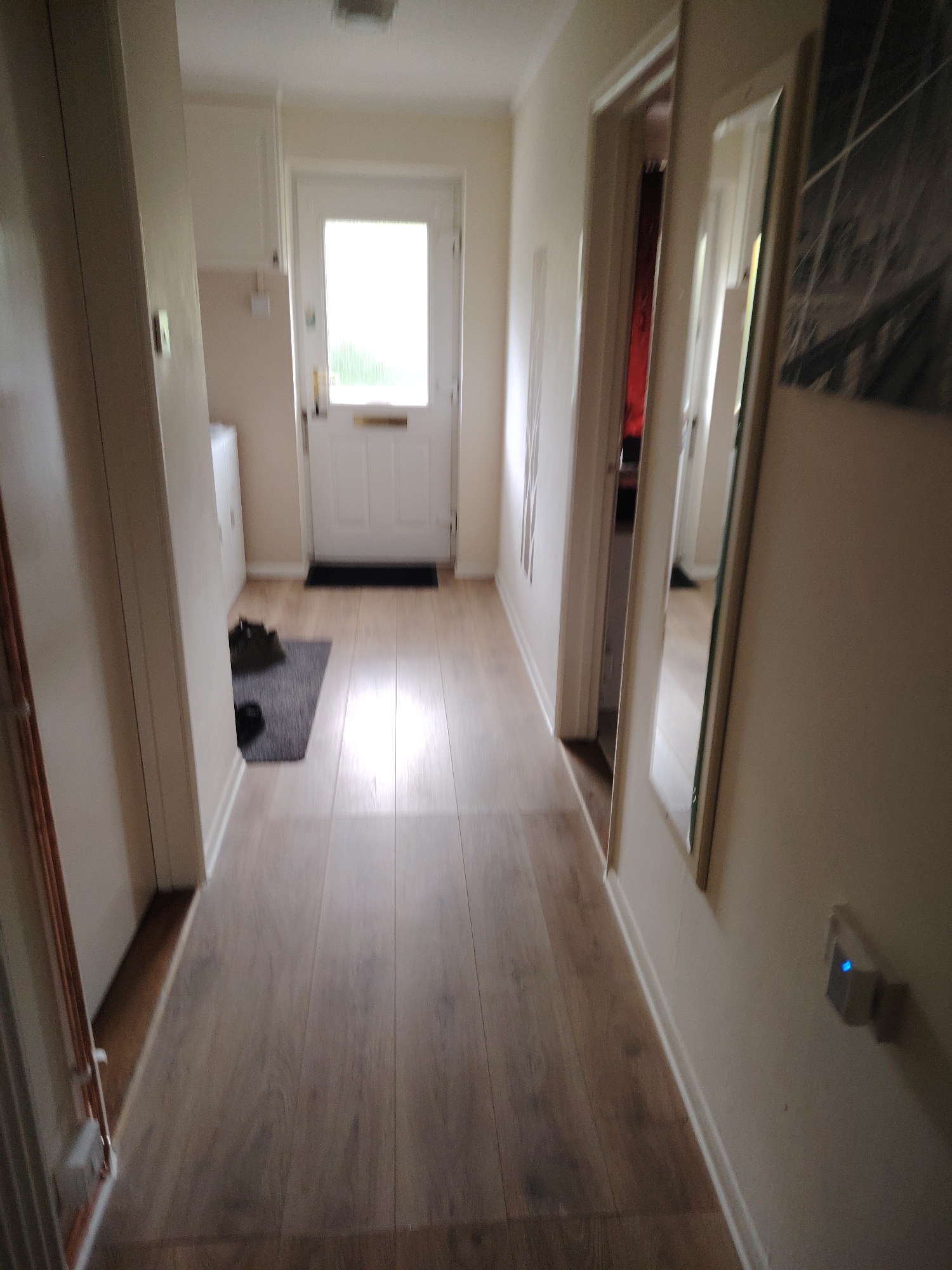 The image captures a residential interior hallway or corridor in Huntingdonshire, United Kingdom, viewed from a slightly elevated perspective looking towards the front of the dwelling. The flooring consists of light-toned, horizontally oriented laminate wood panels that reflect ambient light. The walls are painted a uniform light, neutral color, likely off-white or cream, complemented by white skirting boards along the base of the walls.
On the far left, a section of a light-colored wall partially conceals copper piping running vertically. Further into the midground on the left, a partial view of a white domestic appliance, possibly a washing machine or dryer, is visible below a white wall-mounted cabinet. A dark grey rectangular mat is positioned on the laminate floor, upon which two dark-colored shoes or boots are resting.
At the end of the hallway, a white exterior door with multiple panels is present. The upper half of this door features a translucent glass insert that allows a significant amount of bright natural light to pass through, revealing an obscured view of green foliage outdoors. The door is equipped with a gold-colored letter plate and a corresponding doorknob. A dark rectangular doormat is situated directly in front of this door.
On the right side of the hallway, an open doorway leads into another room, partially revealing a dark red element within that space, possibly a wall or curtain. Adjacent to this doorway, a full-length mirror with a light-colored frame is mounted on the wall, reflecting portions of the opposite side of the hallway and the internal doorway. Closer to the foreground on the right wall, a white electrical socket is visible, into which a small, white, square electronic device is plugged, emitting a distinct blue LED light.
The scene is devoid of any individuals. The lighting is natural, predominantly from the bright door at the hallway's end, creating some areas of direct illumination and shadow.