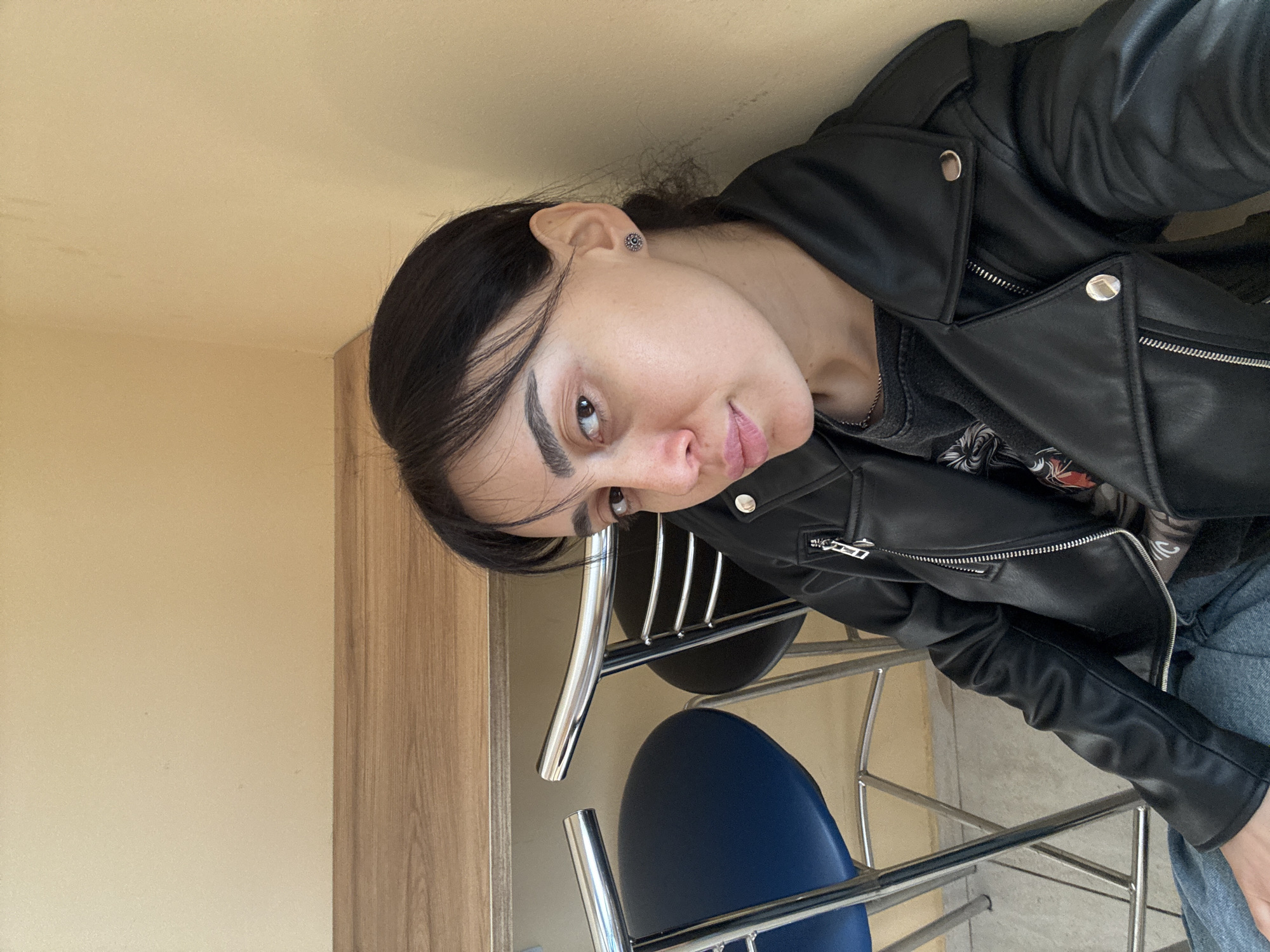 A selfie of a light-skinned person with dark hair, wearing a black leather jacket. The person has dark eyebrows and an earring in their left ear. The background includes a light yellow wall, a wooden cabinet, and a stackable chair with blue seating and a chrome frame. The location could be Tbilisi, Georgia, although there is no explicit marker of location in the image.