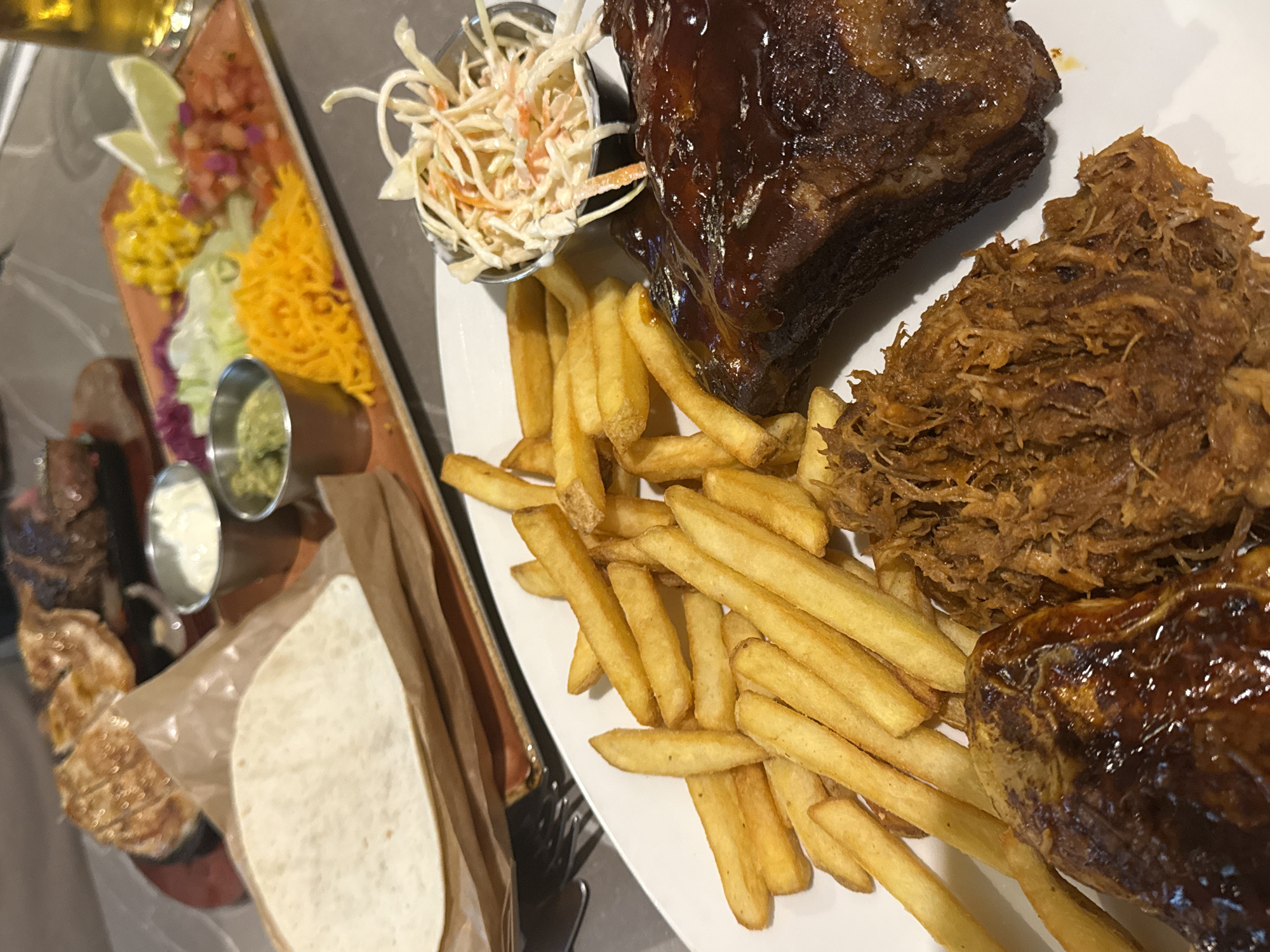 The image displays a large, varied meal presented on a table, likely in a restaurant setting in Milan, Italy. The primary focus is a large white oval plate laden with food, occupying the right side of the frame.
On this plate, a generous portion of golden-brown french fries forms the base. Alongside the fries are two substantial meat components: one appears to be a portion of barbecued ribs or a similar cut of meat, heavily coated in a dark, glossy barbecue sauce. Adjacent to it is a large pile of shredded, slow-cooked meat, resembling pulled pork or beef, with a rich brown color and fibrous texture. A small metal cup of creamy coleslaw, made with shredded cabbage and carrots, is positioned above the sauced meat.
To the left, a rustic wooden serving board holds an array of colorful taco or fajita toppings. These include bright red diced tomatoes (pico de gallo), yellow corn kernels, light green shredded lettuce, purple shredded cabbage, and a pile of shredded yellow cheese. Several fresh lime wedges are also arranged on this board. Further down on a smaller wooden board, two small metal cups contain sauces: one with a white, creamy sauce (possibly sour cream or mayonnaise) and another with a green, chunky sauce, likely guacamole or a herb dip. Partially visible in the bottom left are additional pieces of grilled meat, possibly steak or chicken, along with a soft tortilla or flatbread wrapped in brown paper, suggesting a build-your-own taco or fajita setup.
The meal appears abundant and well-prepared, suitable for sharing or a very hungry individual. The diverse components suggest a Tex-Mex or barbecue-style cuisine. The lighting is soft and ambient, indicating an indoor dining environment. No people or visible text are present in the image.