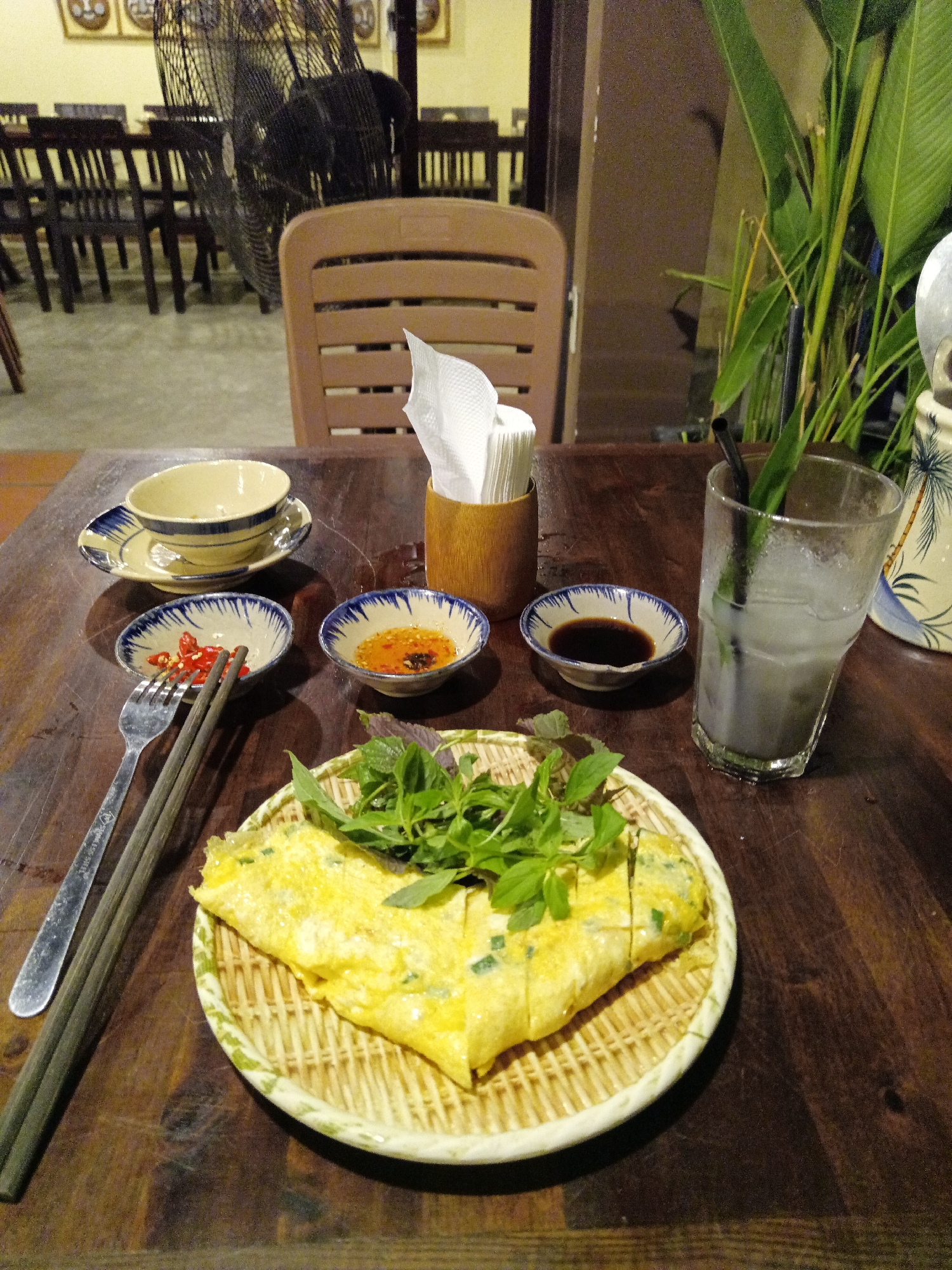 The image depicts a dining table in a restaurant. Located in Nha Trang Ward, Vietnam, the scene is set indoors. On the dark wooden table, there is a woven plate with what appears to be a cut omelet topped with greens. Next to it are chopsticks and a fork, followed by small dishes containing chili peppers, a reddish sauce, and dark soy sauce. There is also a bowl and saucer set. To the right, there is a glass of a beverage with a straw, along with a tissue holder and a vase-like object, possibly a pitcher, with plants. A chair is positioned at the table, and in the background, there are more tables and chairs, along with a large fan and indistinct wall decorations. The room appears to be dimly lit.