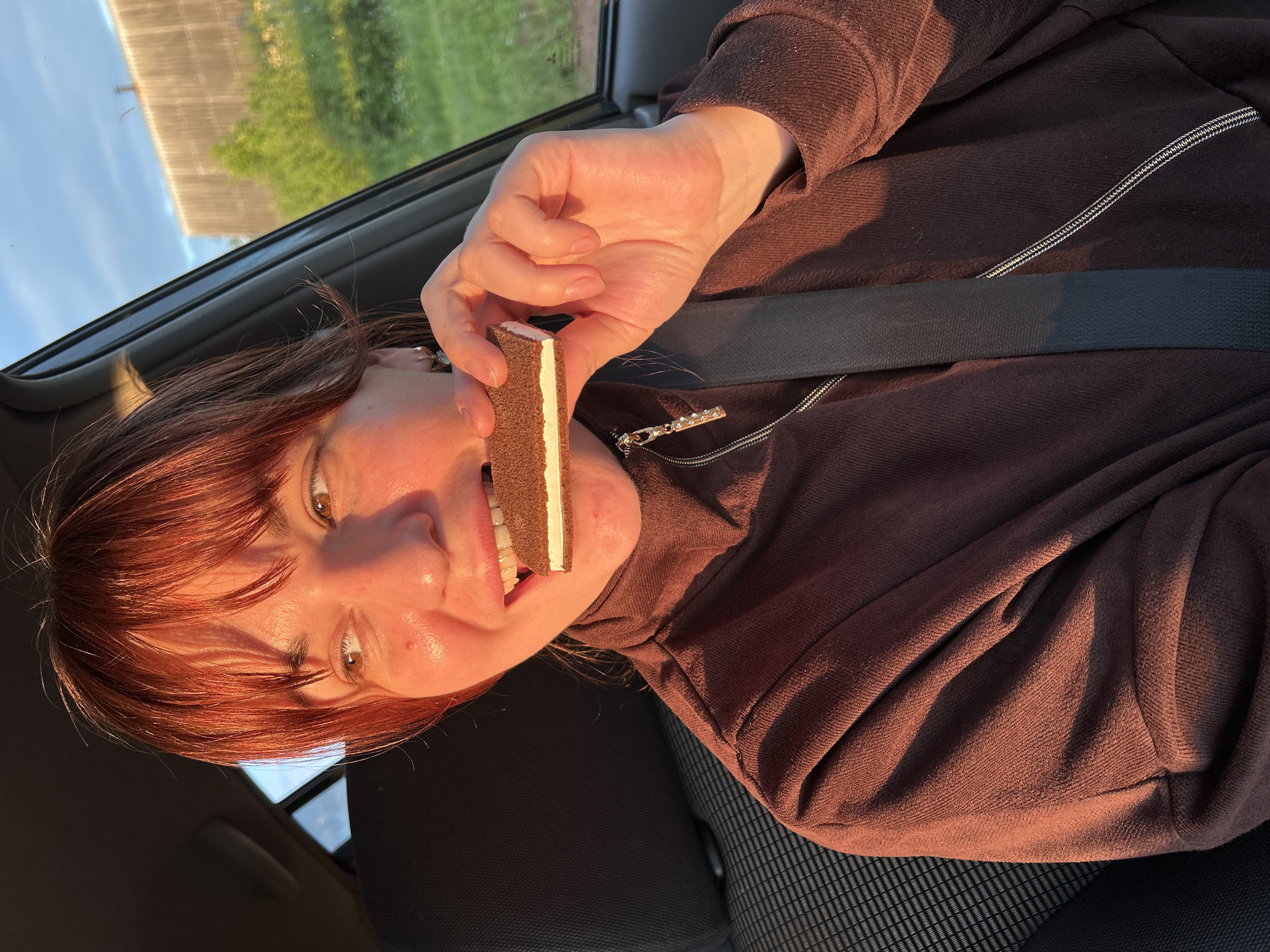 The image features a person (female) with reddish-brown hair and light-colored eyes, positioned in what appears to be the passenger seat of a car. She is captured looking directly at the camera with a playful, smiling expression, her mouth slightly open. She is wearing a brown, long-sleeved top with a zipper, and a dark seatbelt is visible across her chest.
In her right hand, she holds a rectangular ice cream sandwich, which has dark outer layers (likely cookie or cake) and a white creamy or ice cream filling. The sandwich is held close to her mouth, suggesting she is about to eat it.
The setting is a car interior, with a patterned car seat visible. Through the car window, a bright blue sky is discernible above some green foliage and a distant, out-of-focus wooden fence or structure. The scene is bathed in warm, golden sunlight, indicative of either late afternoon or early evening (golden hour), which highlights her features, hair, and clothing. The overall mood is cheerful and casual. There is no visible text in the image. The location context provided is Фоки, Russia.