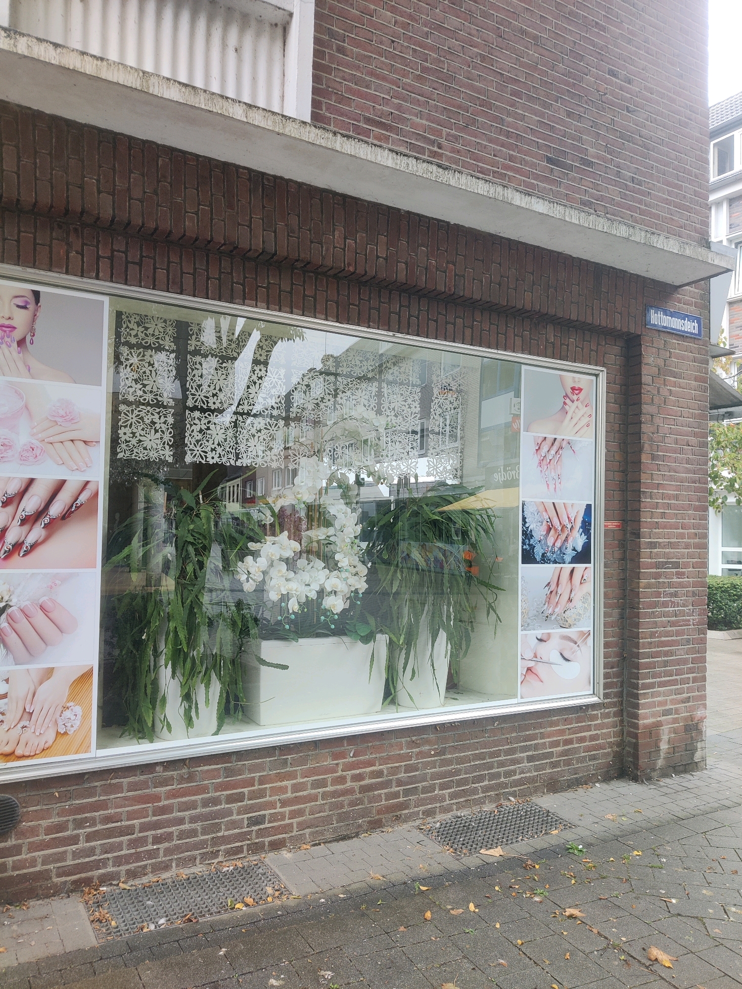 This is a full-frame, eye-level, outdoor photograph of a brick building with a storefront. The storefront has a large glass window displaying advertisements for nail services, including images of manicured hands and feet, and close-ups of nails with elaborate designs. Inside the window, there are several large white planters containing lush green plants, including a large orchid plant with white blooms. The building is located on "Hottomannsweg" in Emmerich am Rhein, Germany, as indicated by a street sign. The sidewalk in front of the storefront is made of grey cobblestones and has a metal grate. Fallen autumn leaves are scattered on the sidewalk and near the grate. The weather appears overcast.