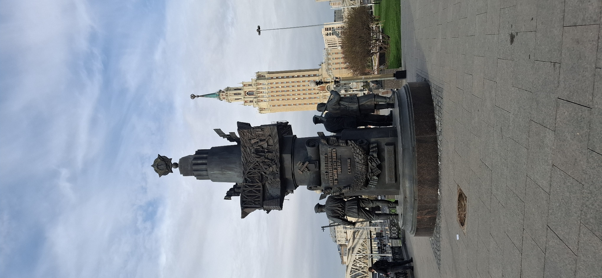 The image shows the Victory Monument in Moscow, Russia, standing prominently against a cloudy sky. The monument, constructed of dark metal, features a tiered design topped with a star. Relief sculptures adorn its sides, depicting scenes of soldiers and warfare. Statues of soldiers stand at its base. In the background, a large building with a spire is visible. The monument sits on a paved area with a sewer drain and a green space with trees.