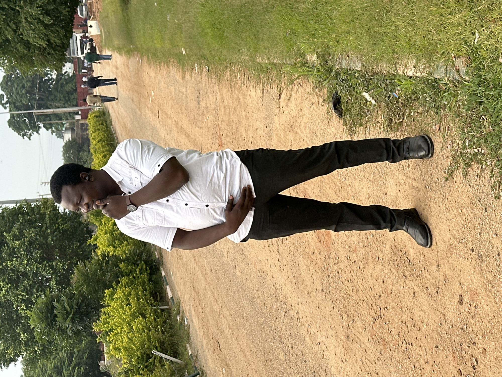 A Black man wearing a white short-sleeved collared shirt and black pants stands on a dirt road in Dadi Riba, Nigeria. He has his right hand on his chin and his left hand on his stomach. He is looking to his right, away from the camera. The road is flanked by green grass on the right and bushes on the left. In the background, several other people are walking on the road, and a white truck is parked further down. Trees and buildings are visible in the distance. The sky is overcast.