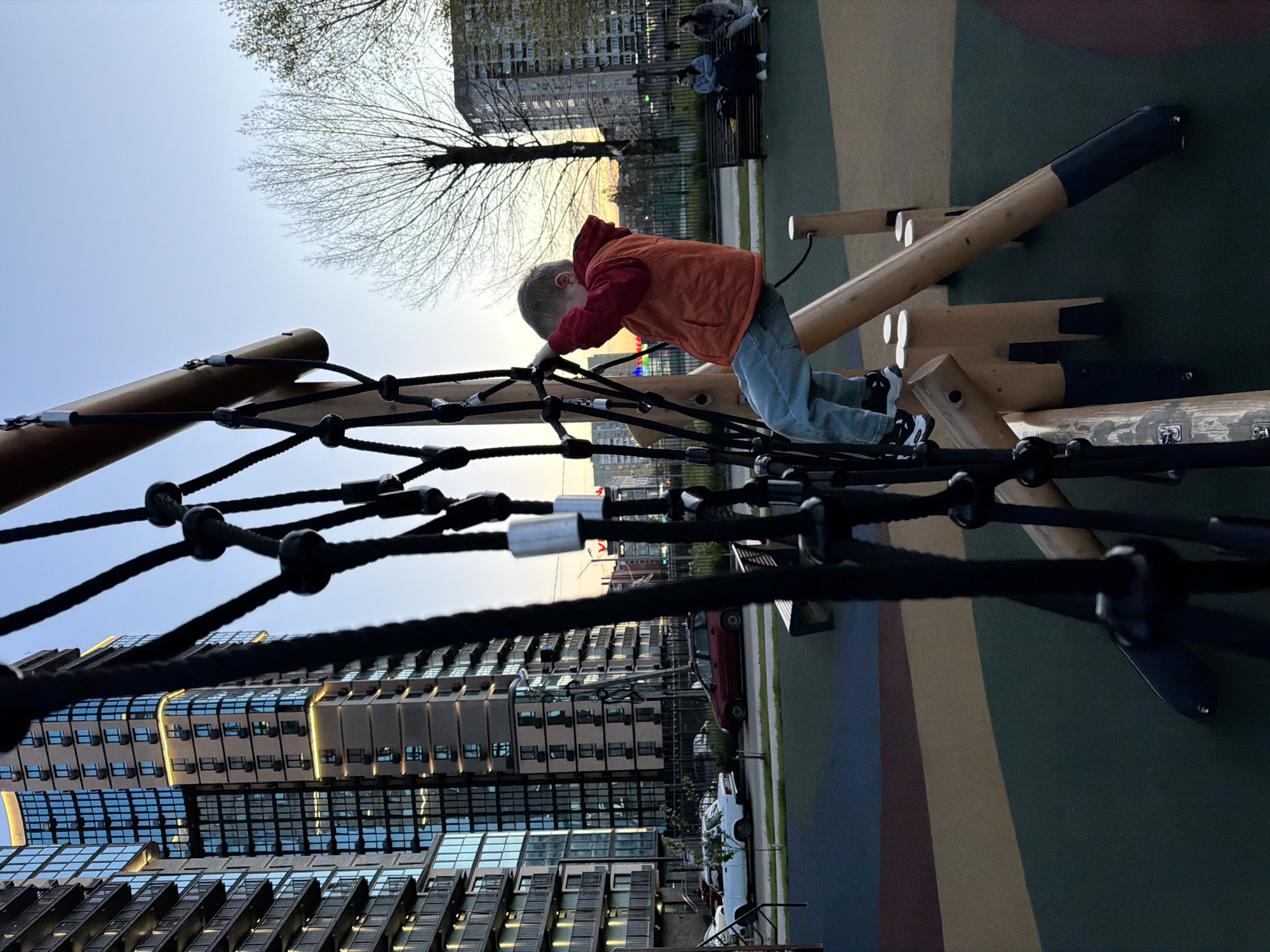 A young child, wearing an orange hooded jacket and light blue jeans, is actively climbing a black rope net structure on a playground. The child is seen from behind, positioned on hands and knees, seemingly focused on navigating the ropes. The climbing structure is made of sturdy wooden beams and thick black ropes. The ground of the playground features a soft-fall surface with patches of green, brown, and dark blue.
In the background, against a clear, light blue sky, several modern, multi-story buildings with numerous windows rise vertically, some reflecting the ambient light. Bare trees, indicative of cooler weather like late autumn or early spring, are interspersed among the buildings and appear against the sky. A soft, warm light on the horizon suggests either late afternoon or early evening, possibly around sunset.
To the right of the climbing child, a small park-like area is visible with a bench where two individuals are seated, and another person is partially seen nearby. Parked vehicles, including a red car and a white one, are visible near the base of the buildings. The scene depicts a calm urban playground setting in Yuzhny, Russia, with the child engaged in play. No text is visible in the image.