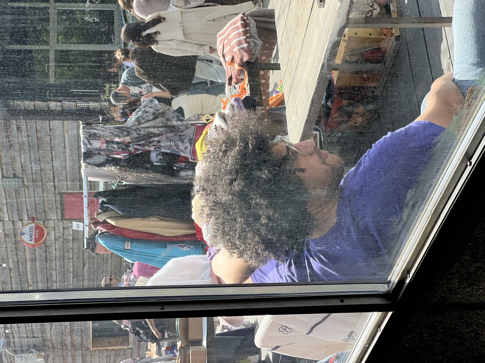 The image, taken from inside a building in Belgrade, Serbia, shows a scene reflected in a window. Through the window, one can observe an outdoor flea market or secondhand clothes sale.
A person with curly hair, glasses, and a beard, wearing a blue shirt, is visible from the neck up, looking towards the flea market outside. They are inside the building, reflected in the window.
Outside, racks of clothes are displayed, along with other items for sale. Several people are browsing through the merchandise. In the background, wooden structures are visible. An “Amstel Bier” sign is attached to the wooden structure.