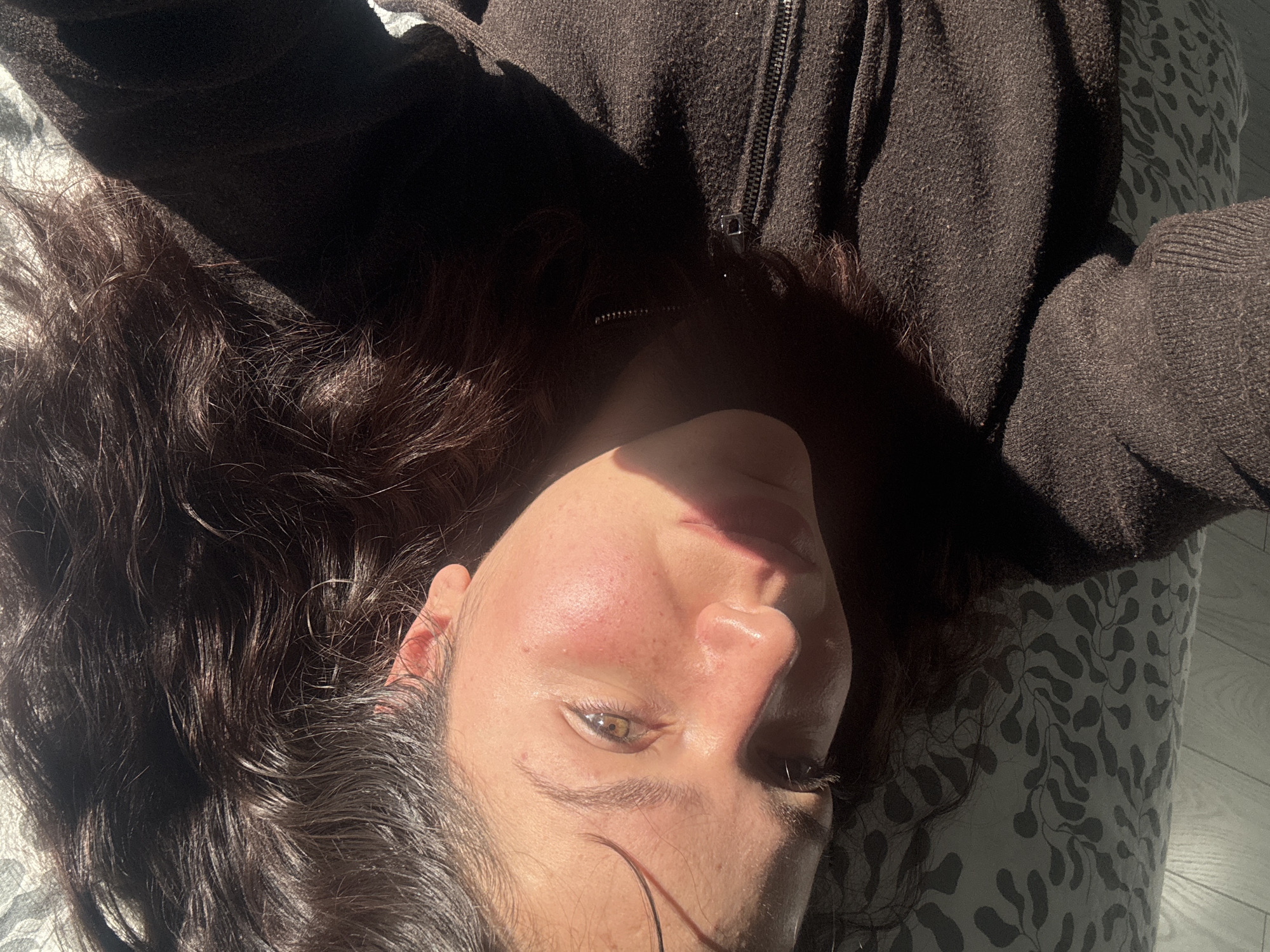 A close-up, high-angle shot depicts a person lying down, looking upwards, with the image rotated so their head is towards the bottom-right. The subject has long, dark, wavy hair spread around their head, partially in shadow and partially catching the sunlight. They are wearing a dark, possibly brown or black, textured hoodie or jacket with a visible zipper down the front.
Strong, directional natural sunlight illuminates parts of their face and hair, creating high contrast with deep shadows. The right side of their face (from the viewer's perspective) is brightly lit, highlighting the cheek, nose, and lips, which appear glossy. The visible eye, which looks directly towards the camera, is light-colored, possibly hazel or green. Some redness is visible on the cheeks, possibly from sun exposure or a natural flush. The rest of the face and much of the clothing are in shadow.
The person appears to be indoors, possibly relaxing on a bed or sofa. The background includes a patterned pillowcase or bedding with a grey leafy or organic design, also partially in shadow. A small section of a light-colored floor, possibly wood or tile, is visible to the right, reflecting some sunlight. The strong, direct light suggests it is either morning or late afternoon. There is no visible text in the image.