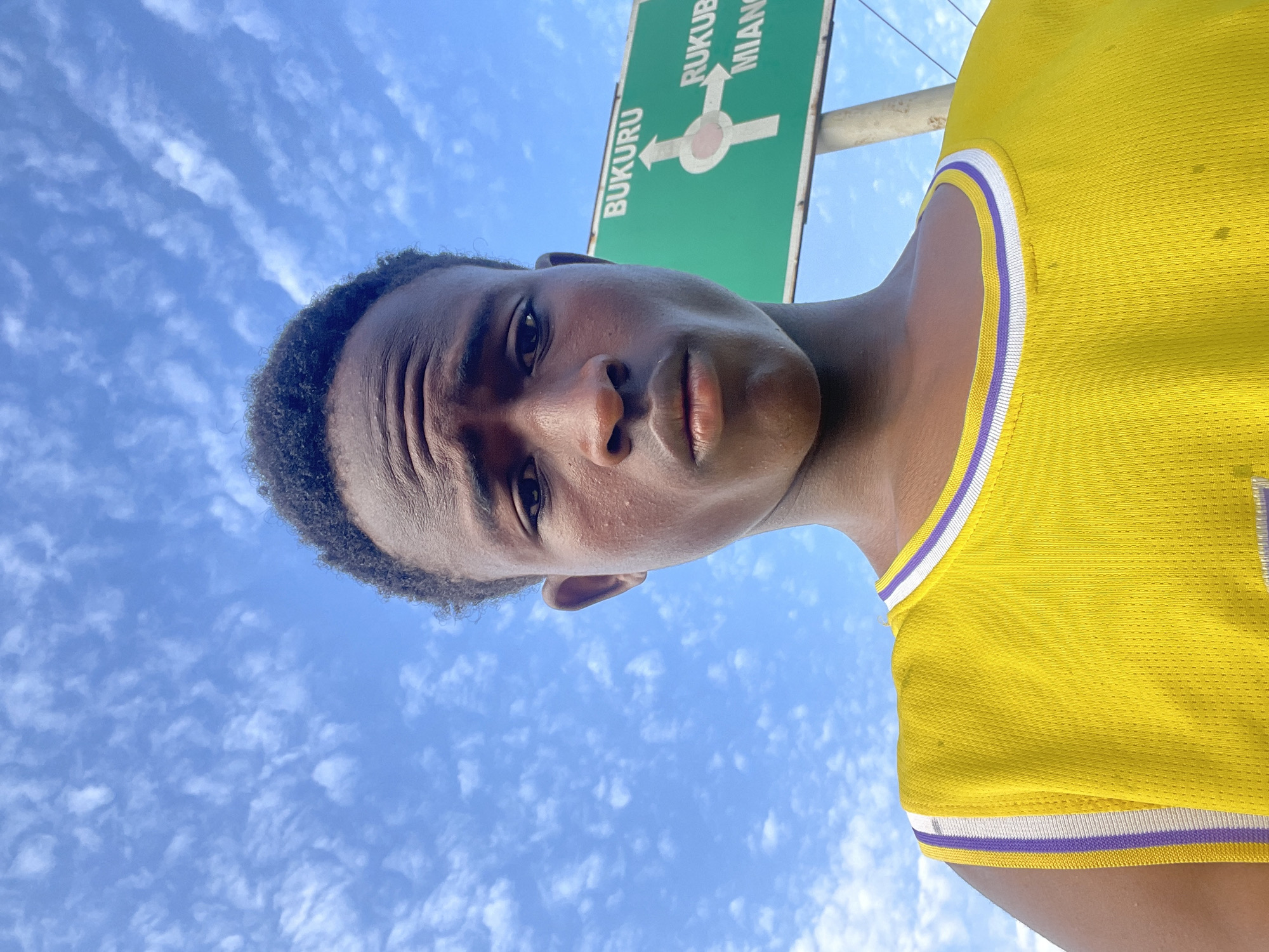 A young Black male is shown from the chest up, taking a selfie outdoors. He has dark, short, curly hair and is wearing a bright yellow basketball jersey with purple and white trim around the neckline and armholes. His expression is serious, and his eyes are looking directly at the camera. Behind him, a green road sign is visible against a blue sky with scattered white clouds. The sign has white text indicating directions, with "BUKURU" at the top and "RUKUBA MIANG" below. A circular directional symbol with arrows is also present on the sign. The scene suggests an outdoor setting, likely in a city like Jos, Nigeria, given the road sign's text.
