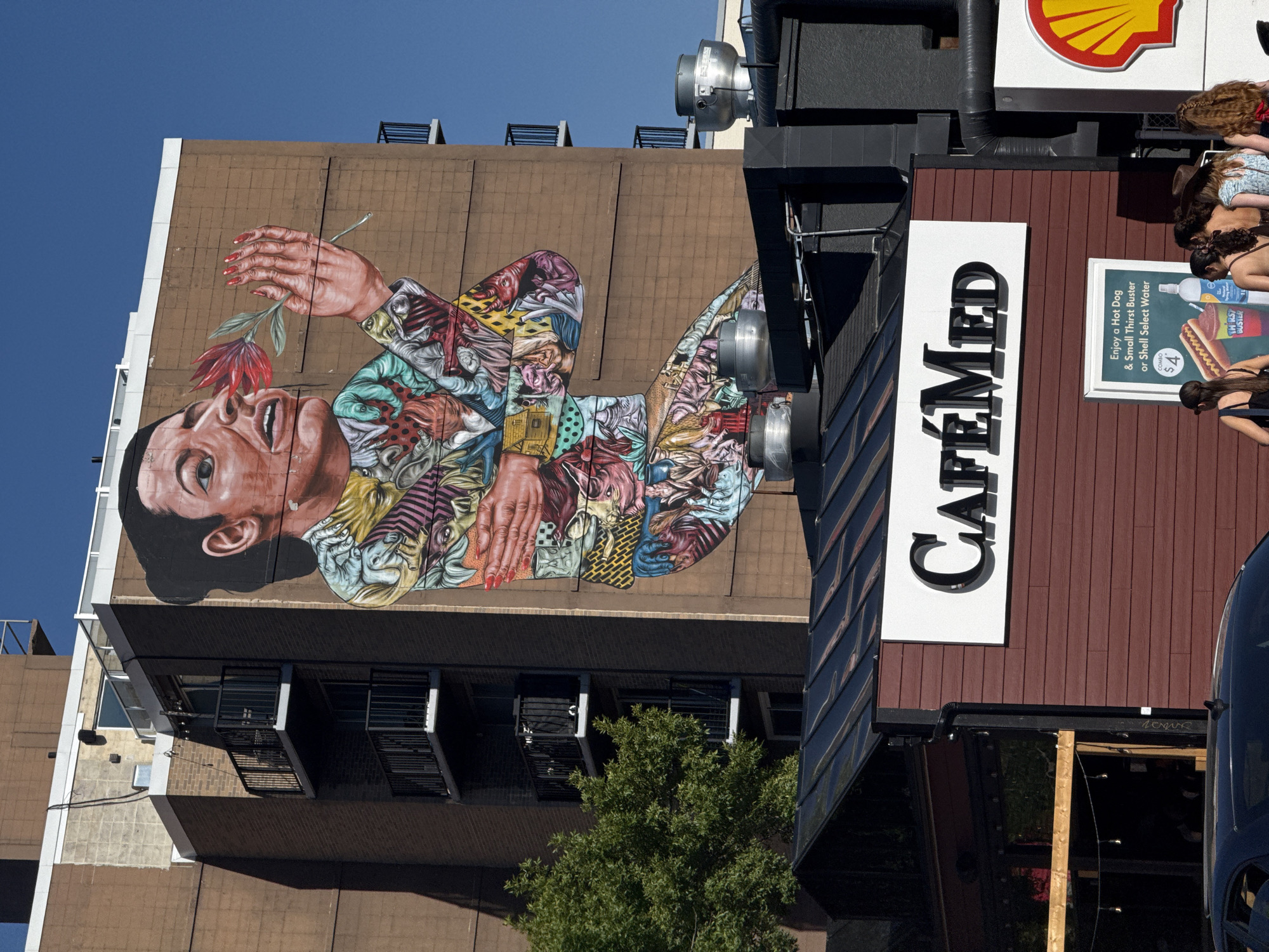 This is a photograph of a mural on the side of a building in Calgary, Canada. The mural depicts a close-up of a woman's face and upper body, with her hand holding a red flower to her nose. The woman's clothing is rendered in a vibrant, abstract style with various patterns and figures. Below the mural, a building with the sign "CaféMed" is visible. In front of CaféMed, there is a poster advertising a hot dog and drink combo. Several people are standing near the poster, facing away from the camera. The sky is clear and blue, and a tree is visible to the left of the mural. The overall scene suggests an urban environment on a sunny day.