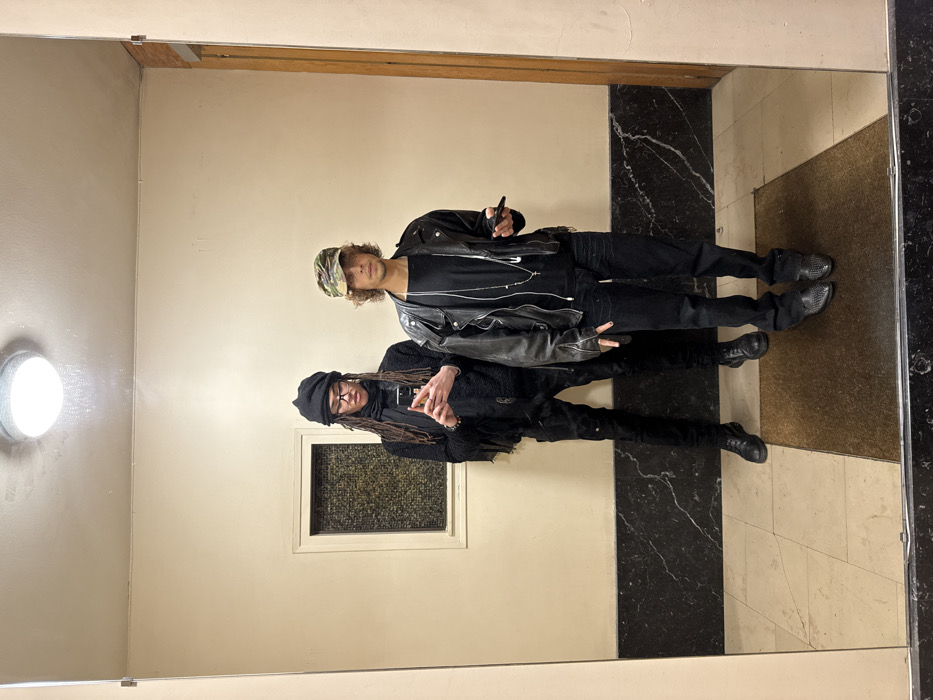 Two individuals are mirrored in a hallway. The one in front wears a black beanie and glasses, dressed in black clothing with dreadlocks. Behind, the other sports a patterned cap, leather jacket over a black shirt, and dark pants. Both hold phones. The hallway features neutral-toned walls, dark marble accents, and tiled flooring. A window is visible to the left of the mirrored reflection. A light fixture adorns the wall near the ceiling.