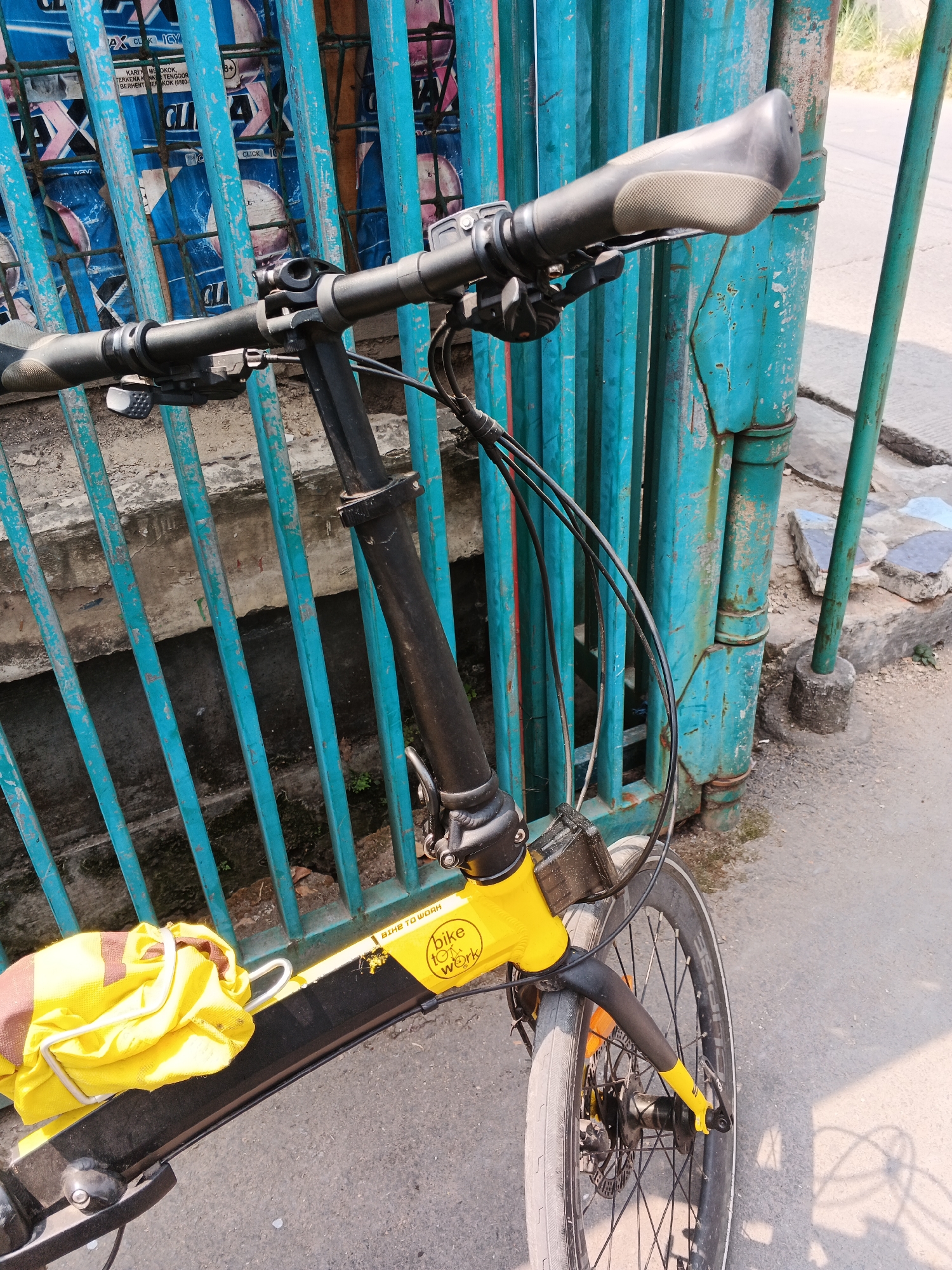 The image shows a yellow bicycle with a brown and yellow bag attached to it, parked in front of a turquoise metal fence in Gandasari, Indonesia. The bicycle has black handlebars with grey grips and is partially visible from the front wheel to the handlebars. The fence appears old and weathered, with peeling paint. Behind the fence, there are posters visible, some of which have blue and pink designs. The ground is paved with asphalt and there is a concrete curb. The lighting suggests it is daytime with shadows cast on the ground.