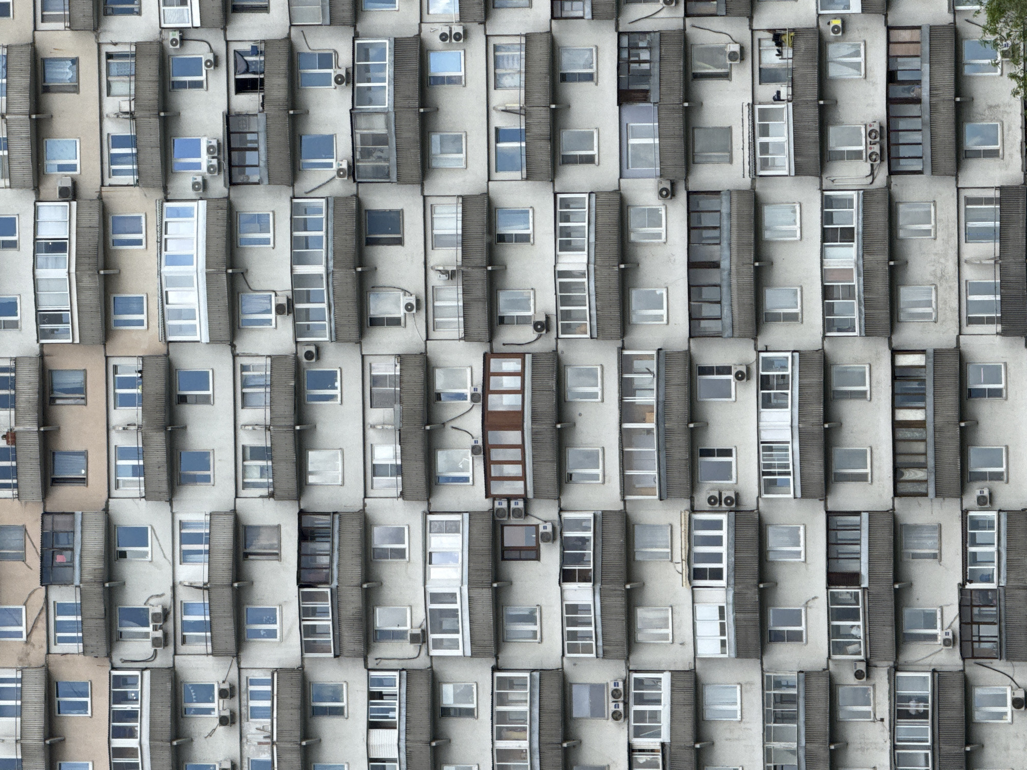 An overhead shot shows the facade of a residential building, likely located in Moscow, Russia. The building is constructed of light-colored, prefabricated concrete panels, punctuated by repetitive arrays of windows. The windows are predominantly rectangular, and some have visible air conditioning units installed near them. Vertical concrete columns separate the window bays. One notable bay has a brown, grid-like design that contrasts with the rest of the facade. A tree branch is visible in the upper-right corner of the frame. The scene is devoid of people. The style of the building suggests a modernist or Soviet-era architectural design.