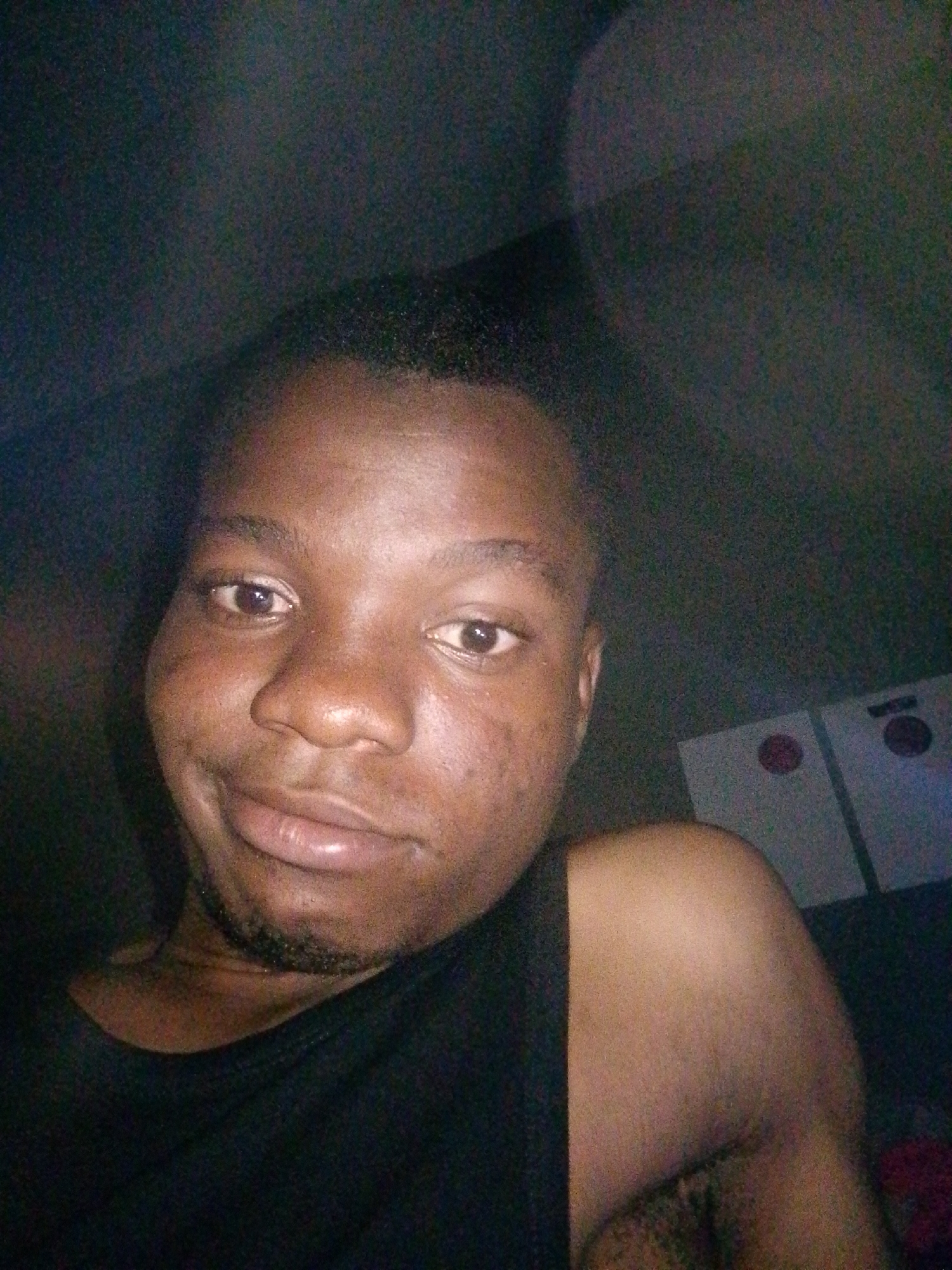 A close-up, dimly lit selfie shows a young Black man wearing a black tank top. He has short hair and is looking towards the camera with a slight smile. His face is turned slightly to his left. In the background, two cornhole boards with red circles are visible. The lighting is low, creating shadows and a grainy appearance.