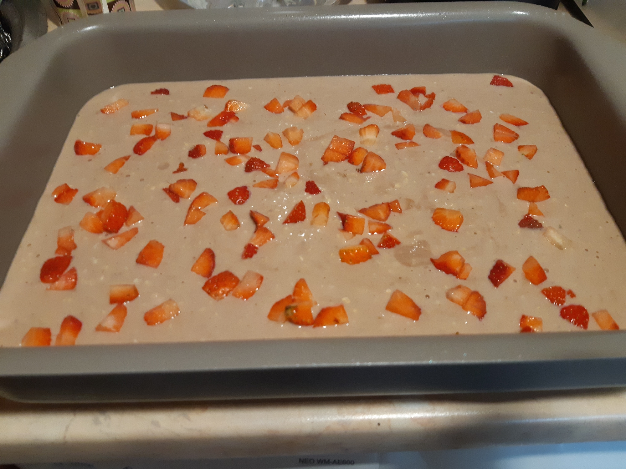 The image shows a gray baking pan filled with batter, sprinkled with diced strawberries. The pan is sitting on a light-colored countertop, with a partial view of a kitchen counter. There is no indication in the image of the location in Plovdiv, Bulgaria, or of any people, actions, or interactions.