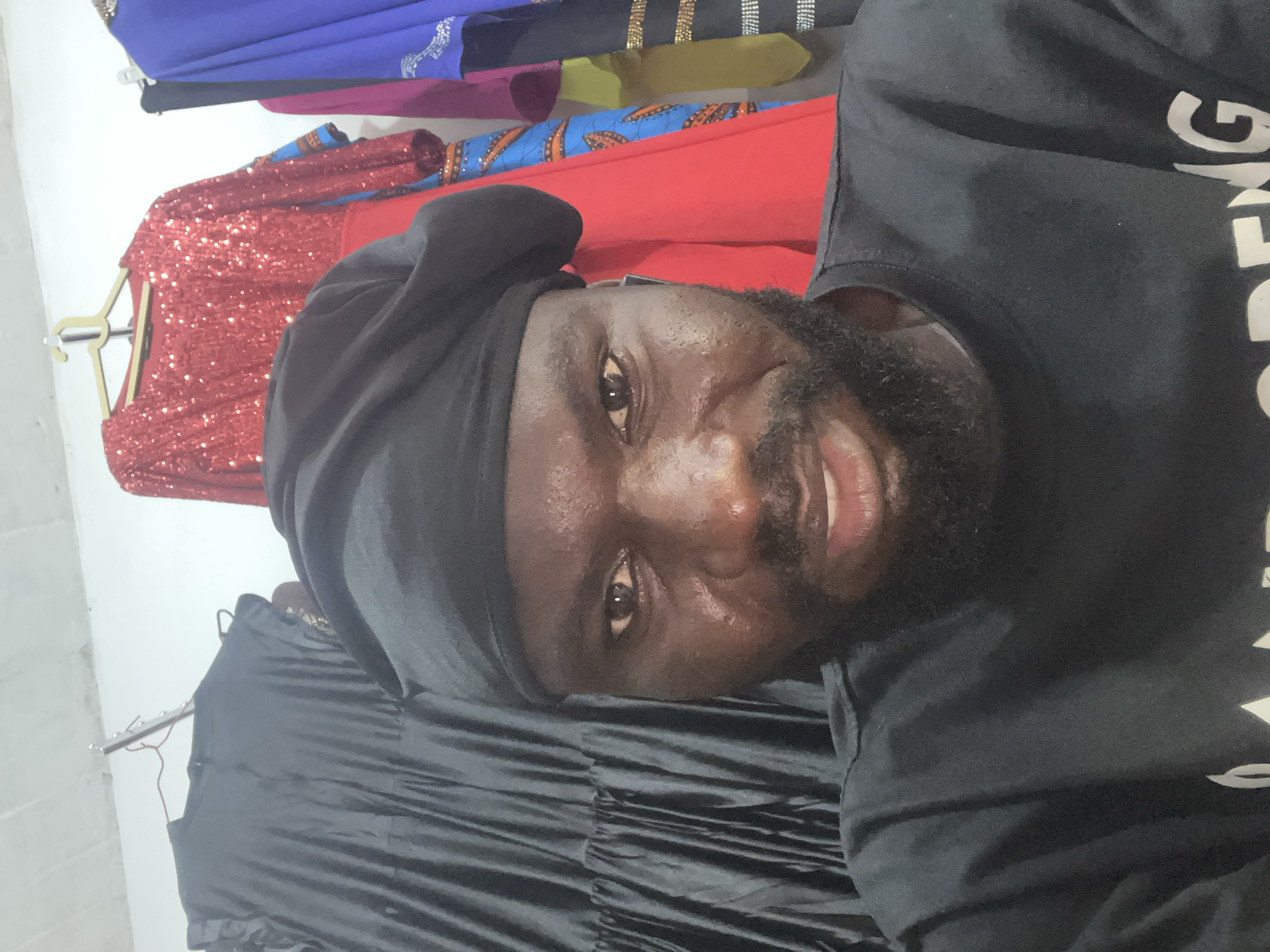 A close-up, tilted shot features a man with dark skin, a beard, and a black head covering. He is wearing a black shirt with white lettering, partially visible. Behind him, clothing hangs on hangers against a white wall. These items include a red sequined dress, a red and blue patterned garment, and other colorful clothes. The image suggests an indoor environment, possibly a shop or storage area, located in the city of Gura Top, Nigeria.