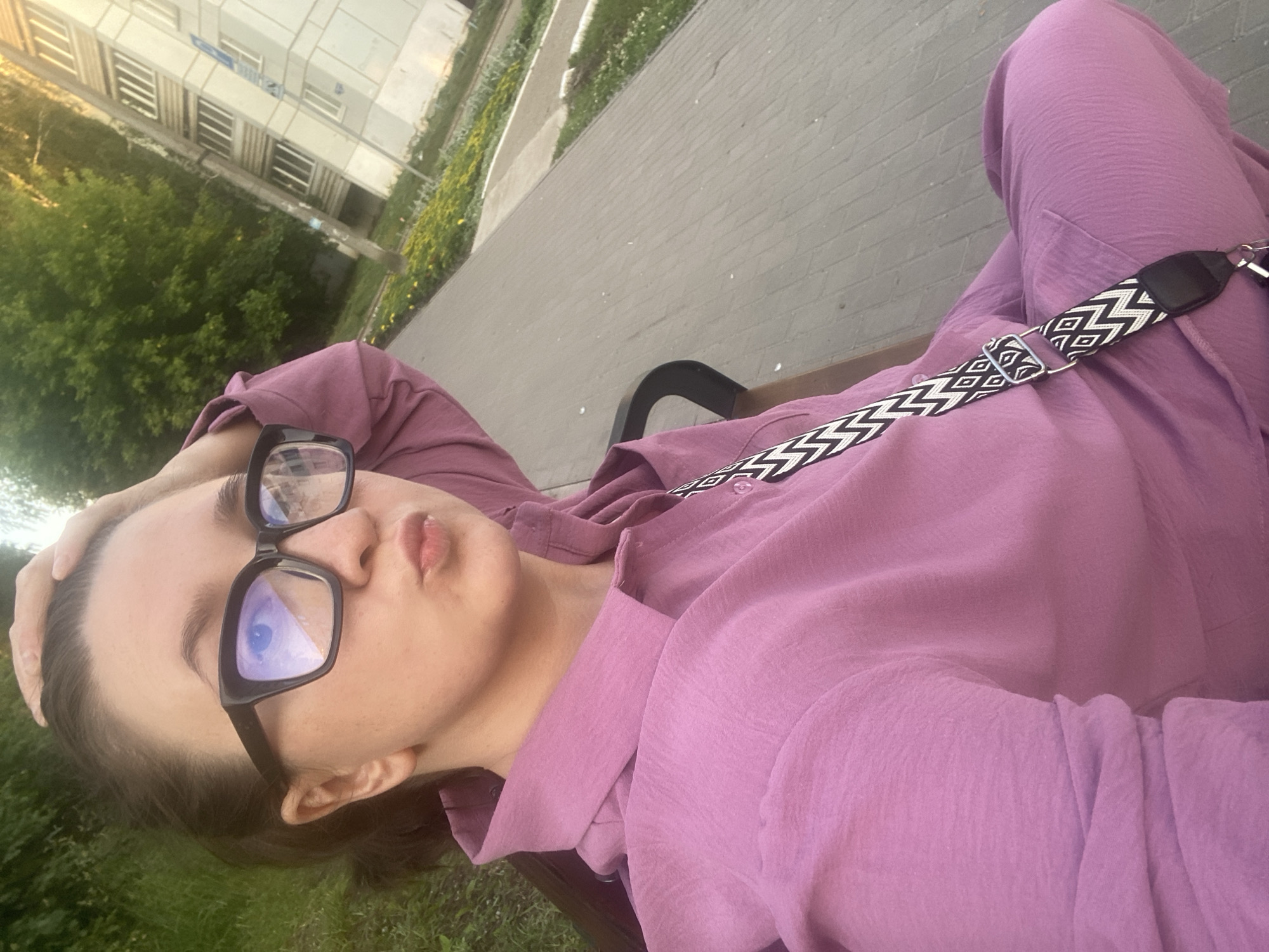 A young woman with dark hair pulled back is sitting on a park bench in Chaikovsky, Russia. She is wearing large black-framed glasses and a pink collared shirt. She is pouting with her lips pursed and has her left hand on the back of her head. A patterned strap of a bag is visible across her chest. The background shows a paved walkway, green grass, trees, and a multi-story building, suggesting an urban park or residential area. The lighting indicates it is likely late afternoon or early evening, given the warm glow on the building and trees.