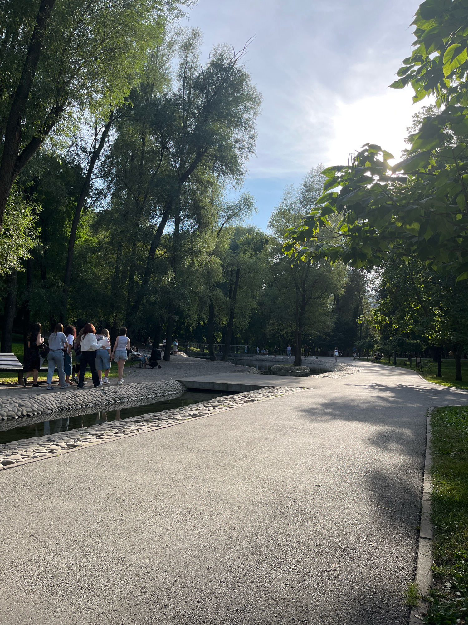 A group of young people walk along a paved path in a park. The path curves to the right, with a grassy area on the right side and a decorative stone-lined water feature on the left. In the background, lush green trees create a dense canopy, with sunlight filtering through the leaves. The sky is partly cloudy with the sun visible. Some individuals are seen further down the path, and there is a stroller visible. The scene appears to be a serene summer day in a city park.