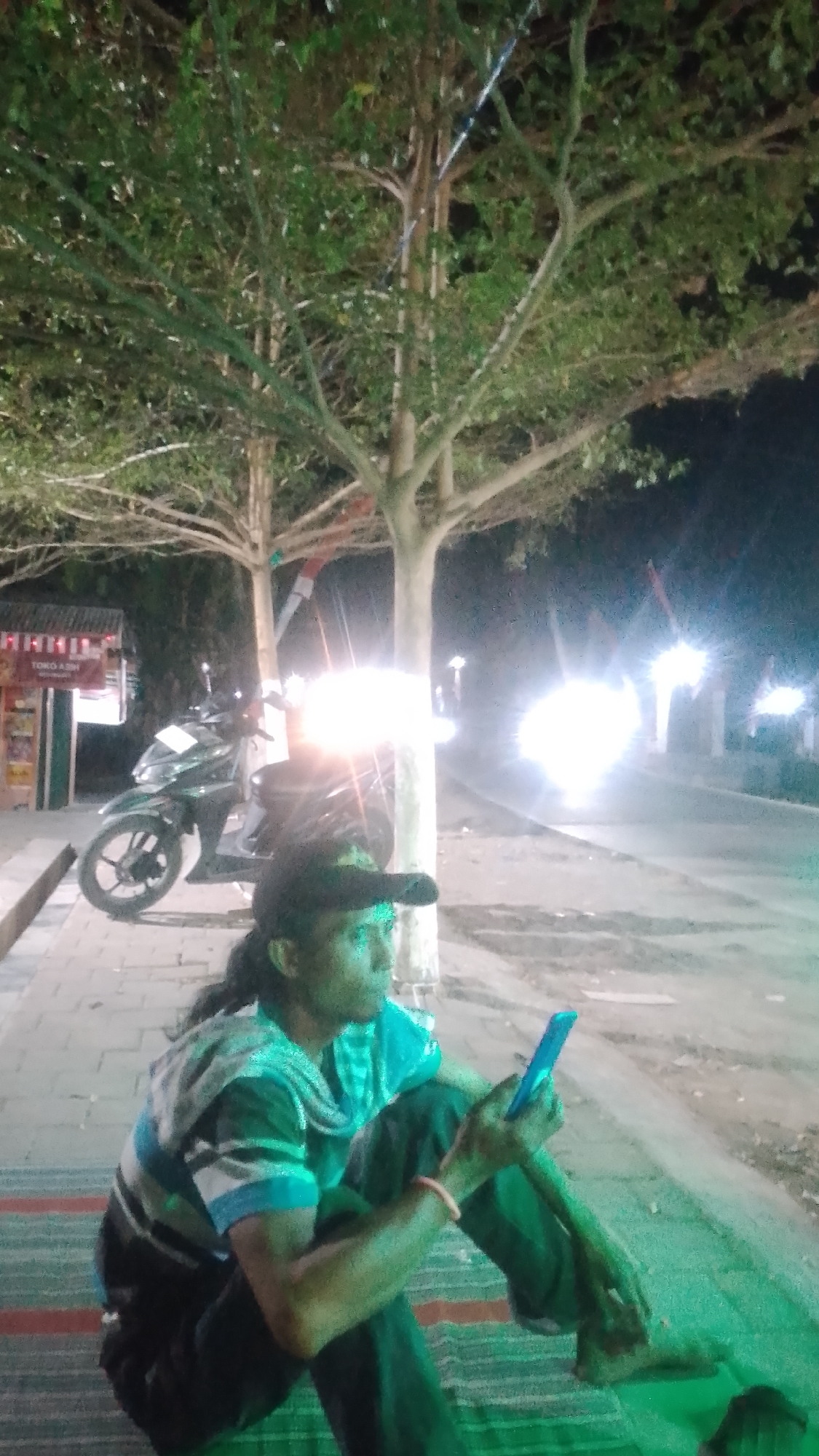 A man wearing a black cap and a striped shirt sits on the ground, looking at his phone. He is in front of a scooter, with trees and a road in the background. The scene appears to be in Karanglangu, Indonesia, as indicated by a sign that says "Toko Asih". The lighting suggests it is nighttime, with streetlights and headlights from vehicles illuminating the scene. The man's attire and the presence of the scooter suggest a casual, urban setting.