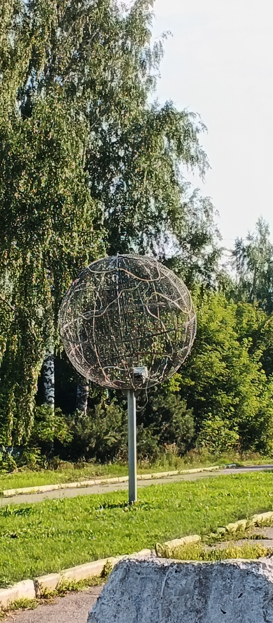 A close-up, eye-level shot shows a large, spherical wire sculpture mounted on a pole in what appears to be a park or green space in the city of Podgornoye, Russia. The sculpture is composed of a dense network of thin, metallic wires forming an open, spherical shape. A small, box-like object, possibly a camera or light fixture, is attached to the pole just below the sculpture. The background is dominated by lush green trees and foliage, with a clear sky visible above. In the foreground, a portion of a large, rough-textured concrete object is visible, along with a paved area and grass. The scene suggests an outdoor public space.