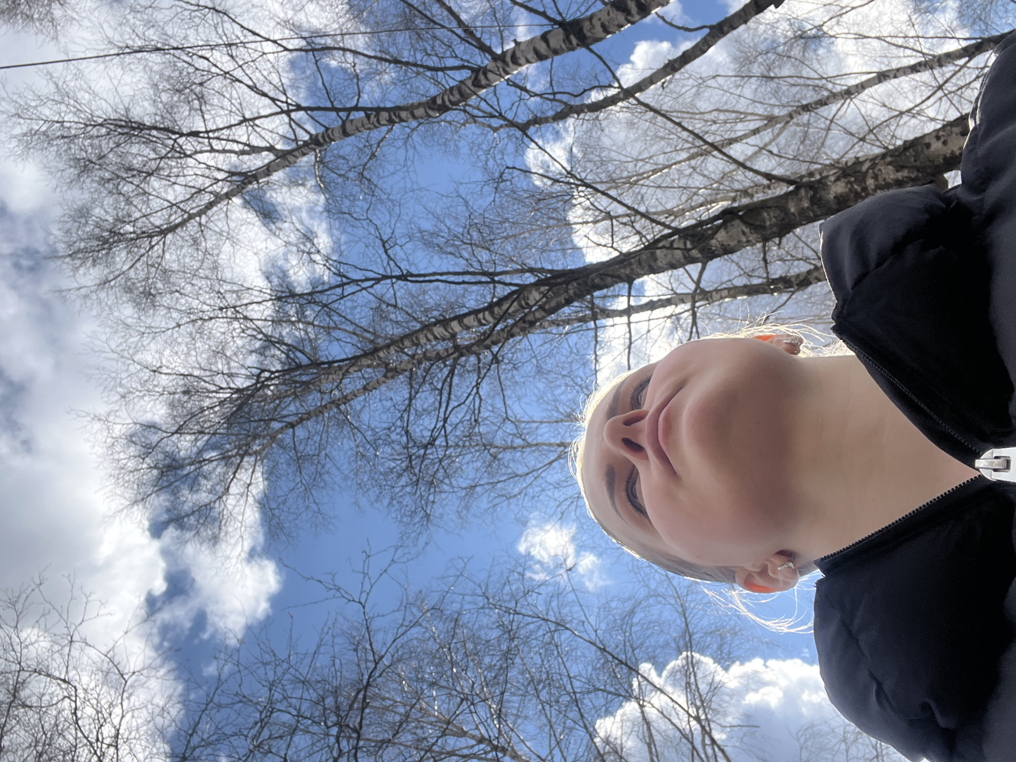 The image is an upward-angled, low-perspective selfie, primarily capturing a person's face against a backdrop of a bright blue sky with scattered white clouds and bare tree branches. The person's face is in the lower right portion of the frame, tilted back, showing the nose, chin, and part of the neck. They are wearing a dark, possibly black, puffy jacket with the zipper visible. The background consists of a view of the sky and bare tree branches, common in late winter or early spring.
Given the type of trees visible and the sky conditions, it's plausible this scene could be from a park or wooded area in Riga, Latvia, although without further contextual information it is impossible to determine the exact location.