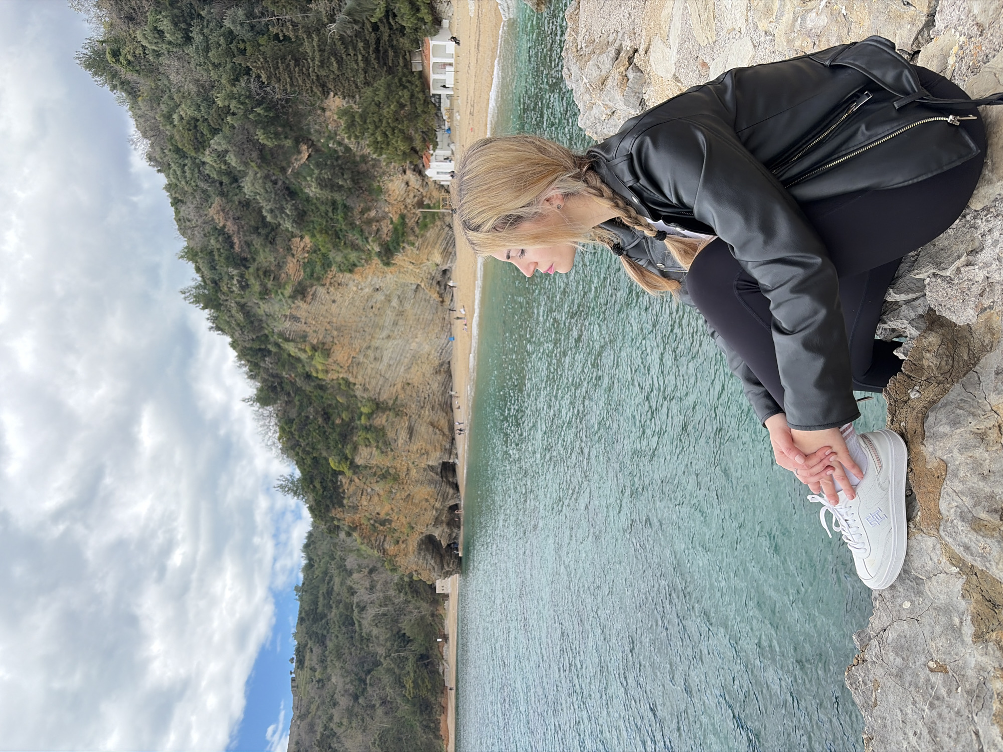 A young, fair-skinned woman with blonde hair in braids, wearing a black leather jacket and black leggings with white sneakers, crouches on a rocky edge overlooking a beach and body of water. She appears to be touching her foot. The location is likely near Budva, Montenegro, as suggested by the coastal landscape including a beach, cliffside with vegetation, and clear blue water. A building is seen on the beach in the distance. The sky is overcast with some clouds.