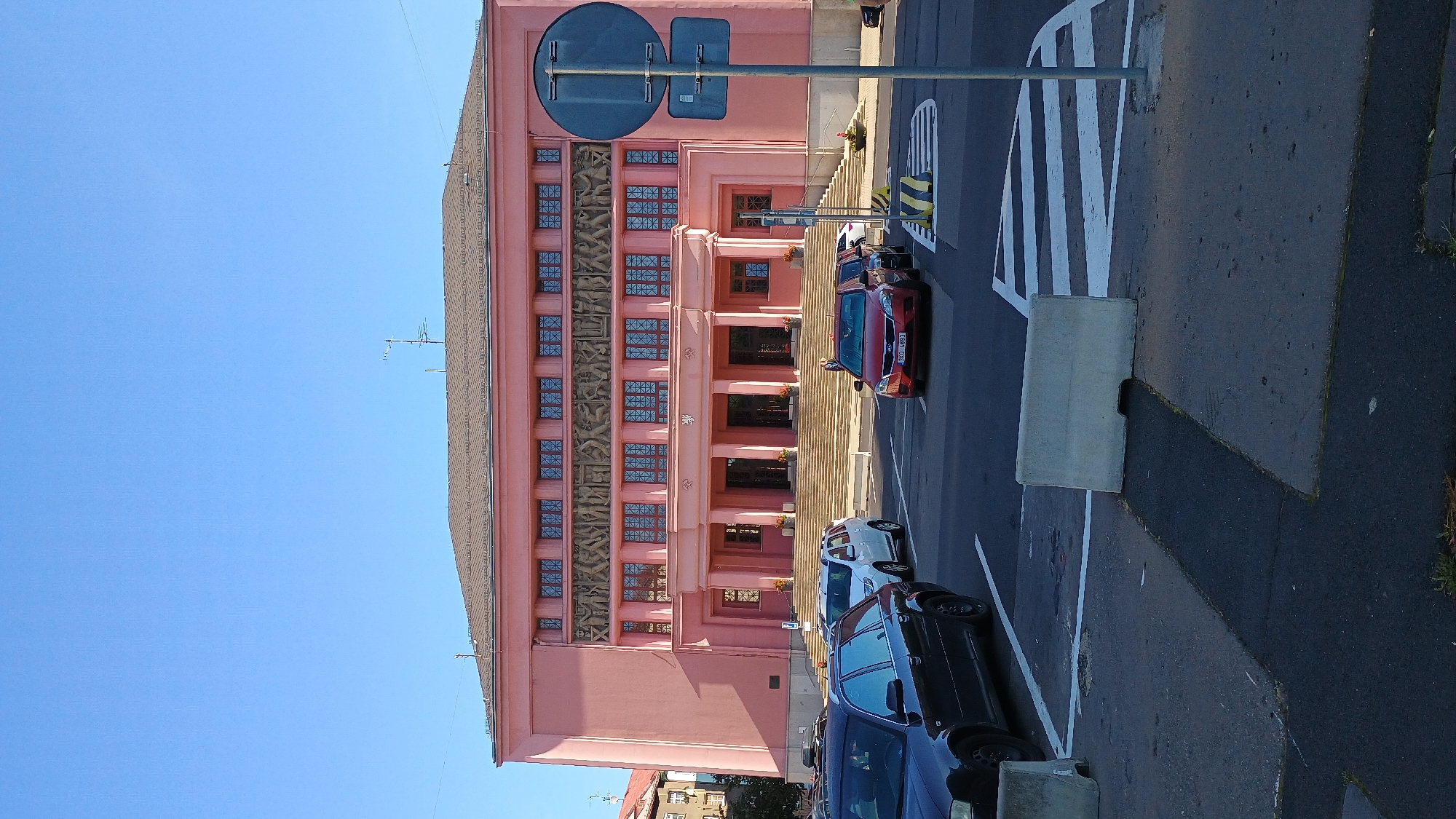 The image shows the Municipal House in Sokolov, Czechia, a pink, neoclassical building with a large, imposing facade. In front of the building, a wide set of stairs leads to a grand entrance. The facade is adorned with a frieze depicting figures in relief. Several cars are parked or driving in the foreground, including a black Audi station wagon, a white SUV, and a red sedan with a license plate reading "DKO 4893". A traffic signpost stands on the right side of the image, and concrete barriers are placed on the road in the foreground, indicating a pedestrian crossing. The sky is clear and blue, suggesting a sunny day.