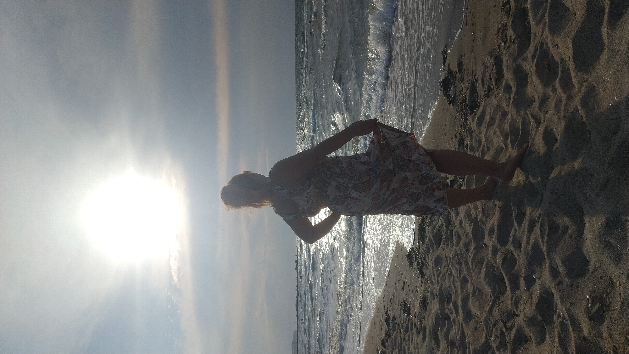 This media file captures a woman standing on a sandy beach in Neoi Epivates, Greece, during what appears to be the late afternoon or early evening, given the low angle of the sun and the warm, bright light. The sun is positioned in the sky to the left of the frame, casting a bright glare and creating long shadows across the sand, indicating a time close to sunset. The weather seems pleasant, with clear skies and calm seas, though there are some light clouds.
The woman, silhouetted against the bright sun and shimmering water, is wearing a patterned dress. She is holding the hem of her dress with her right hand. Her hair is pulled back, and her pose suggests a moment of reflection or enjoyment of the seaside. The foreground is dominated by the textured sand of the beach, showing distinct footprints and ripples. The ocean is visible in the middle ground, with gentle waves lapping at the shore. In the distance, a thin, hazy line suggests land or a distant shoreline.
The scene evokes a sense of tranquility and relaxation, typical of a beach setting at the end of the day. There is no visible text in the image.