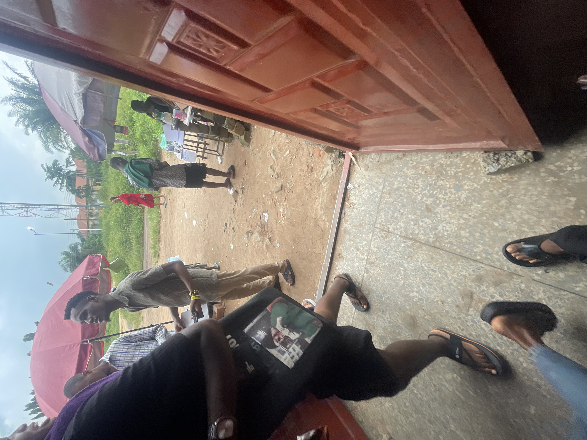 In Ihiagwa, Nigeria, under a cloudy sky, a group of people are gathered outdoors. In the foreground, a person wearing a black t-shirt with a printed image and black shorts is holding a black bag. To their right, a person in khaki pants and a patterned short-sleeved shirt stands, looking upwards. Behind them, a large red umbrella provides shade. Further in the background, a woman in a red dress walks away from a stall with umbrellas, and a person wearing a green backpack is standing near a table. The ground is dirt, with scattered debris. A large, red metal door dominates the right side of the frame, angled towards the viewer. The scene suggests an outdoor market or gathering area.