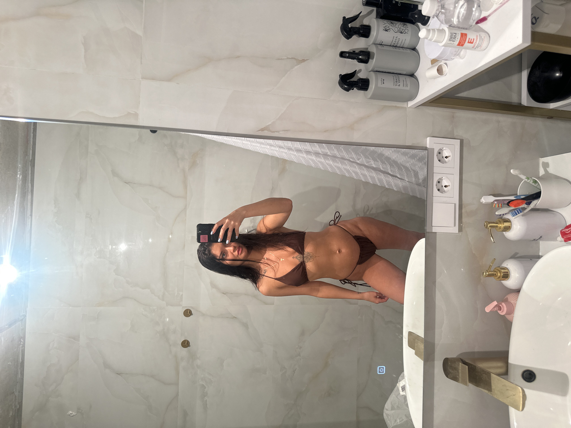 A woman is captured in a mirror selfie in what appears to be a modern bathroom. She has long, dark hair and is wearing a dark brown two-piece bikini. Tattoos, including a mandala-style design, are visible on her lower stomach/hip area and near her collarbone. She holds a black smartphone in her right hand, pointing it towards the mirror to take the photo.
The bathroom features light-colored walls with a marble-like pattern. A large mirror serves as the central element of the shot. To the right of the woman's reflection, a white textured shower curtain is partially visible.
Above and to the right, a white shelf with a gold frame holds various personal care products. These include several grey spray bottles, some labeled "KOKONA GRAND" and "TOBACCO & VANILLA." Other bottles are visible, such as a clear one and a white bottle with "LIBRE erm" and "E ВИТАМИН" (E VITAMIN) printed on it. A black spherical object also rests on the shelf.
Below the shelf, a white sink with a modern golden faucet is present. A white cup holds multiple toothbrushes next to soap dispensers, one partially showing "Colgate." Two white European-style electrical outlets are mounted on the wall. A glowing golden power icon is visible on the bottom right of the mirror's surface, suggesting it is an illuminated mirror. The setting appears clean and well-maintained, consistent with a contemporary indoor bathroom in Moscow, Russia.