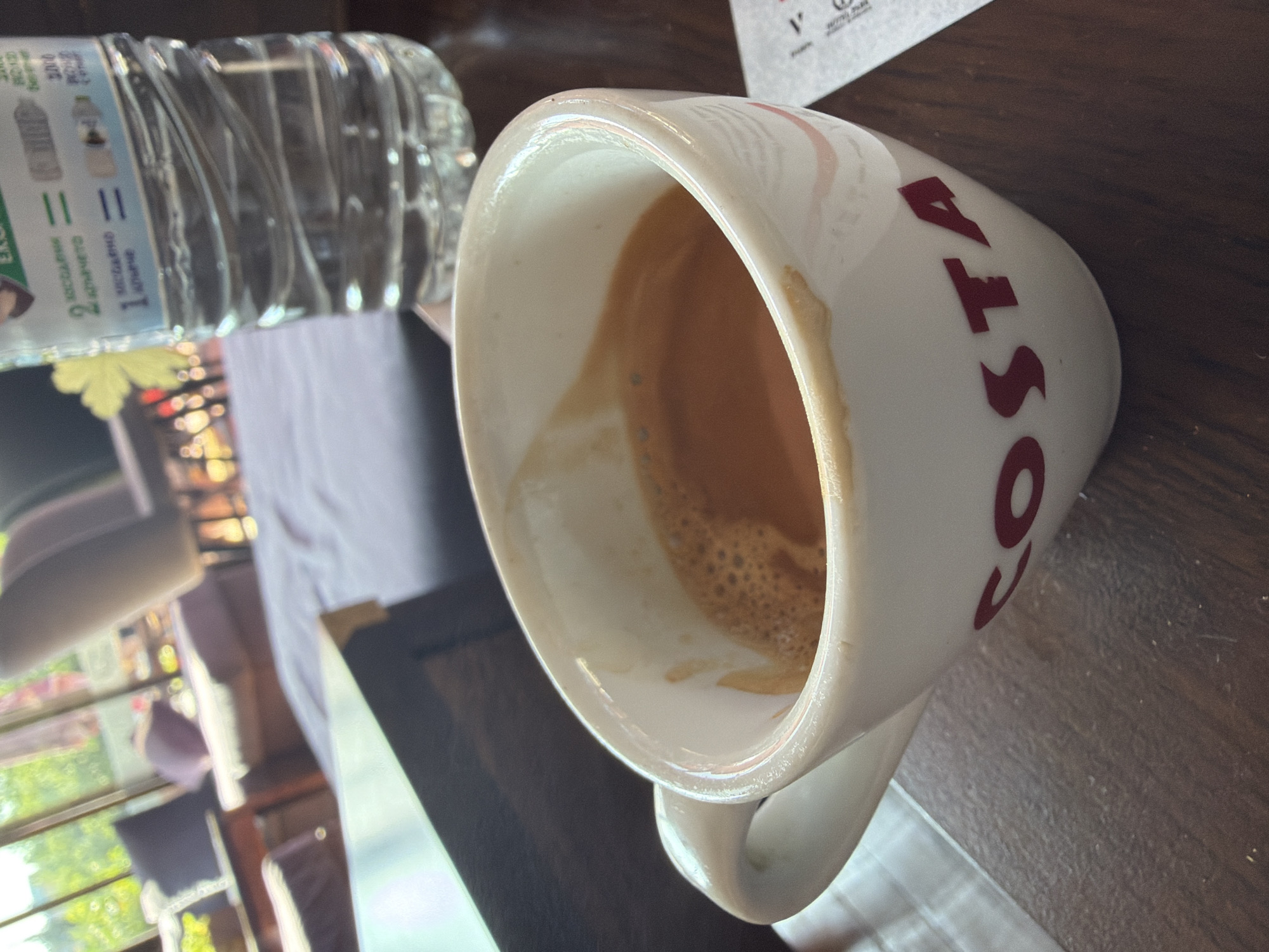 The image captures a white ceramic Costa coffee mug resting on its side on a dark wooden table. Inside the mug, a small quantity of residual coffee, complete with crema and foam, is visible. The iconic red "COSTA" logo is prominently displayed on the mug's exterior. Adjacent to the mug, a clear plastic water bottle lies horizontally, with parts of its label visible. The label contains Cyrillic text, including "ЕКСТРАКТ" (Extract), "КОНЦЕНТРАТ" (Concentrate), and "БЪЛГАРИЯ" (Bulgaria), along with the numbers "2" and "1", suggesting details about the water's content or origin, consistent with the location context of Blagoevgrad, Bulgaria.
The background is softly blurred, indicating a shallow depth of field. This blurred area reveals bright daylight streaming from a window, and indistinct shapes of indoor furniture such as chairs and tables, suggesting a cafe or restaurant setting. The scene portrays a calm, daytime moment, possibly after a coffee has been consumed. No individuals are visible in the frame.