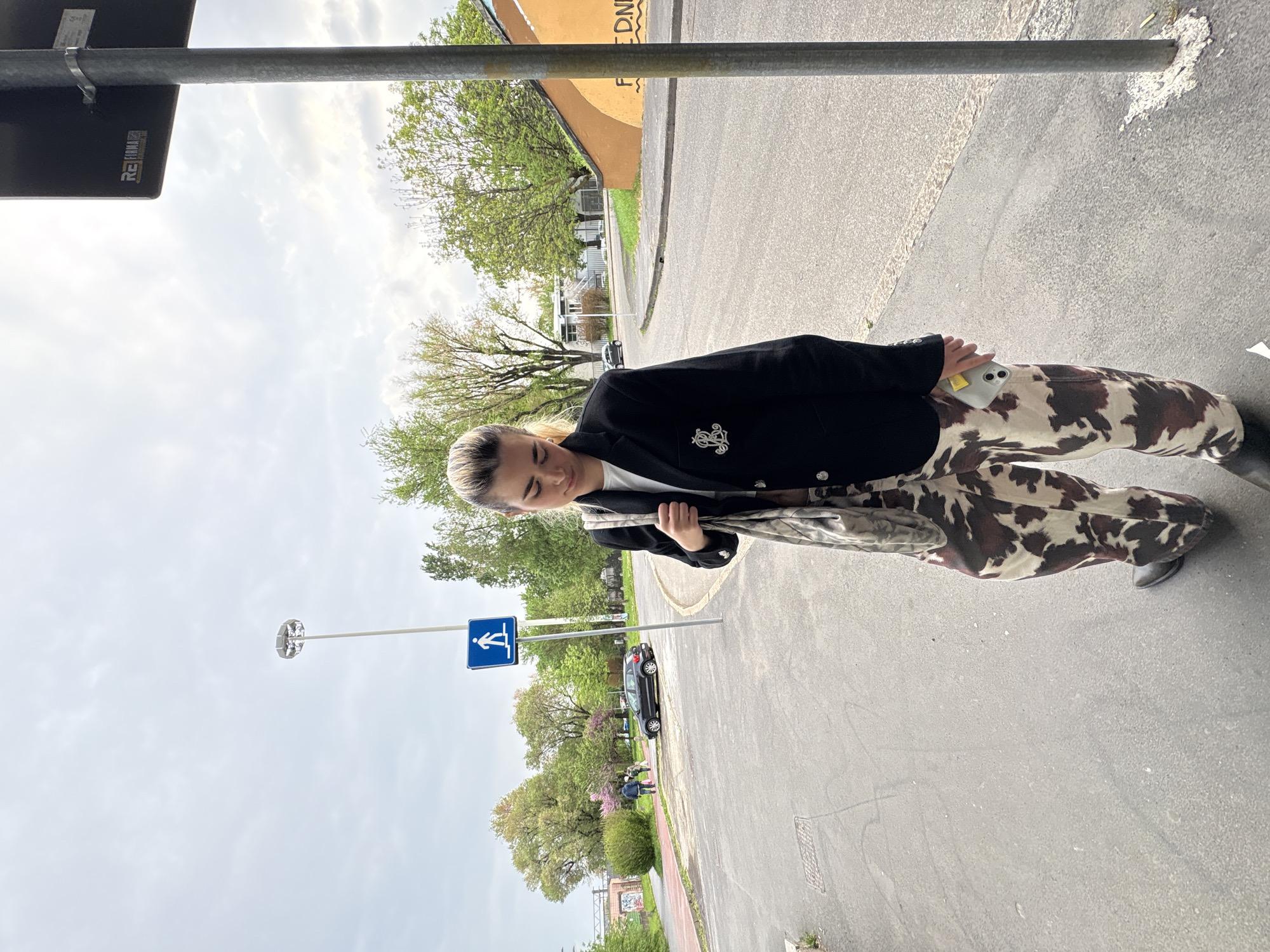 A person stands on a paved sidewalk, likely in a European city, given the road markings and signage. The individual wears a black blazer with an emblem on the lapel, a white shirt underneath, and brown and white cow-print pants. They are holding a phone and a camo-patterned bag. A street sign with a pedestrian crossing symbol is visible in the background, along with a car parked on the street. The background includes buildings and trees. The photo is taken in daylight under cloudy conditions. There is no evidence to definitively confirm the location as Milan, Italy, without additional context or information, but given the street signs and architecture, it is likely a European city.