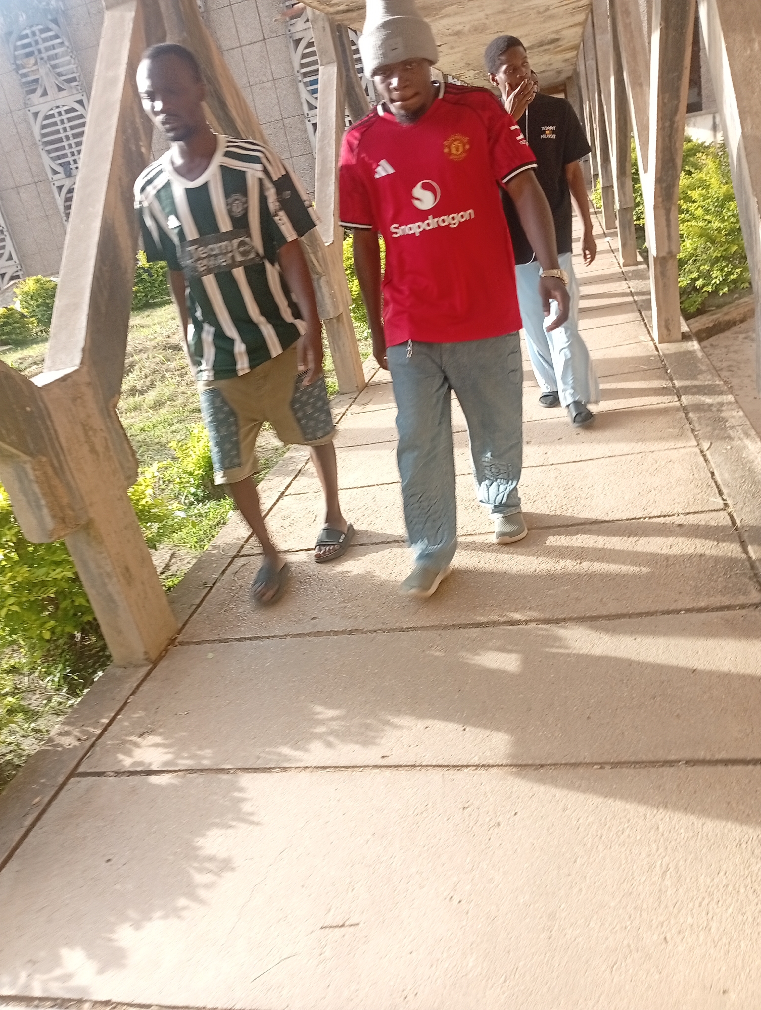 Three young men walk along a concrete pathway bordered by grassy areas and vegetation. The man in the foreground on the left wears a dark green and white striped shirt and khaki shorts. The man in the middle wears a red Manchester United jersey with a white collar and light blue jeans. The man in the background on the right wears a black t-shirt and light blue pants. The pathway is covered in dappled sunlight and shadows cast by overhead structures. The architecture suggests an outdoor walkway with visible concrete pillars and a patterned ceiling or overhang. The setting appears to be a public space.